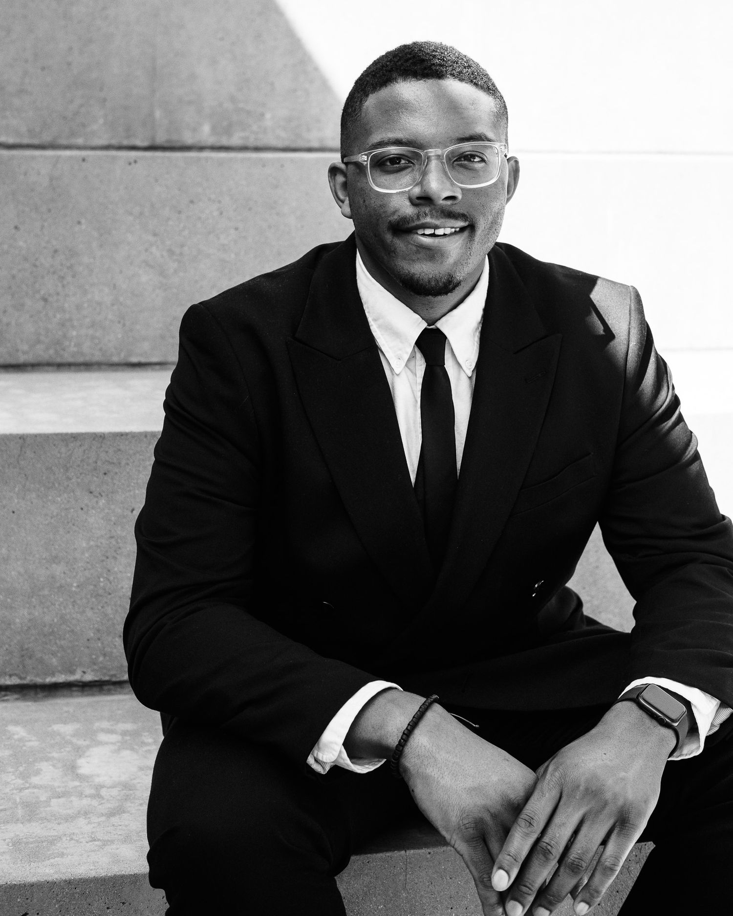 Black and white portrait of a smiling man in a suit and glasses sitting on steps.
