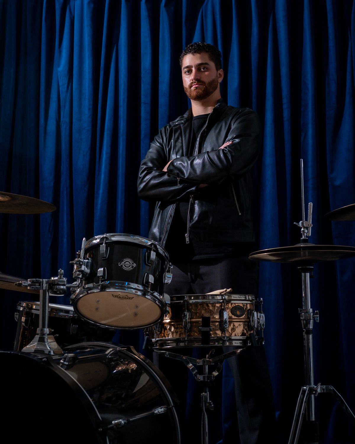 A bearded drummer in a black leather jacket stands confidently with arms crossed behind a drum kit against a blue curtain.