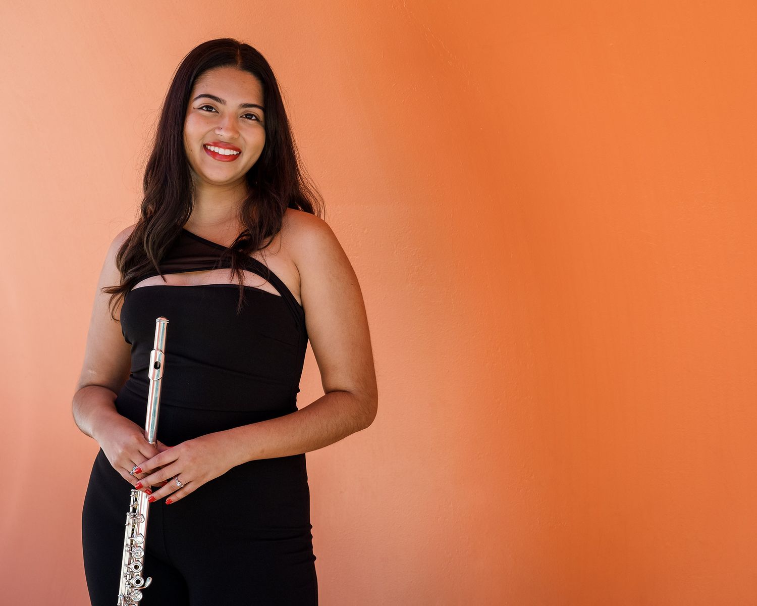 A smiling young woman in a black dress holds a silver flute against a bright orange background.
