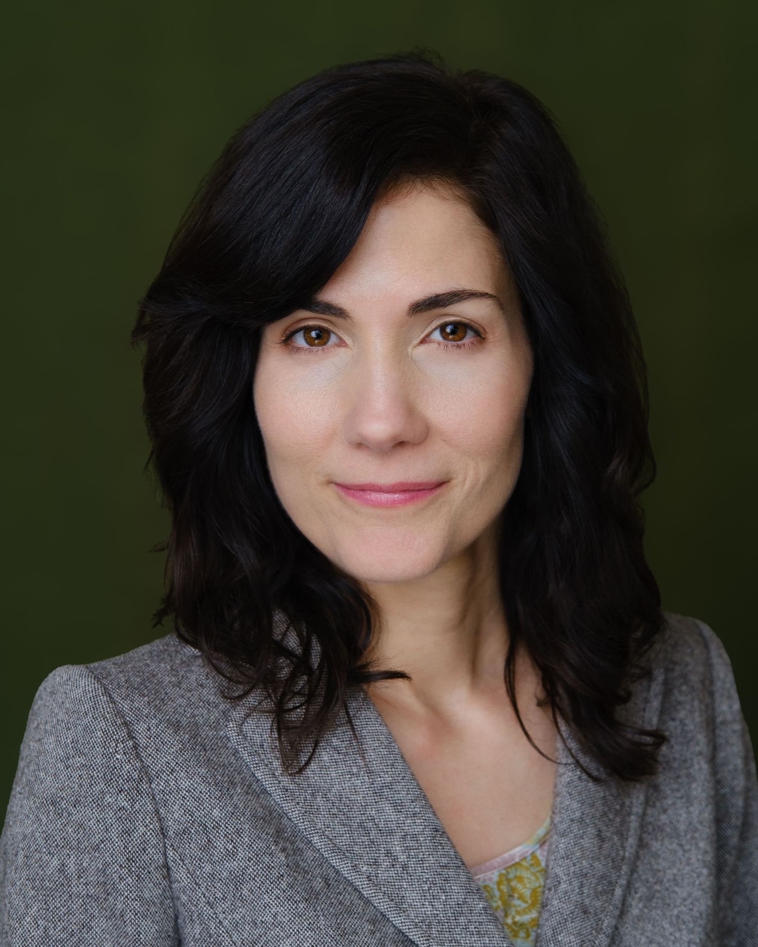 Professional headshot of a woman with dark wavy hair wearing a gray blazer against a green background.