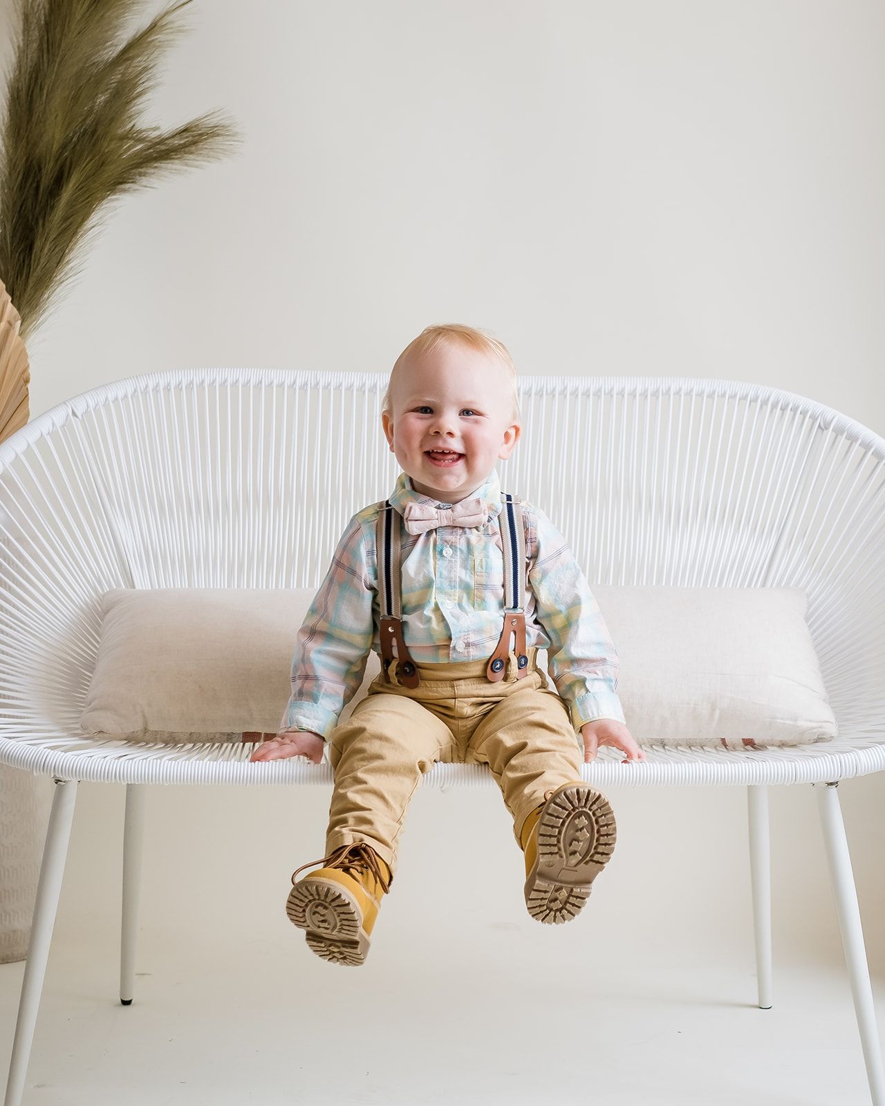 Happy toddler boy in suspenders and bow tie sitting on a white wicker bench smiling.