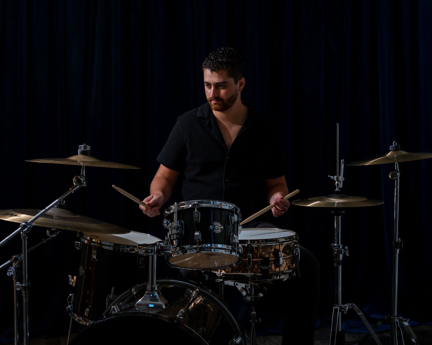 Drummer playing a drum kit on a dark stage under dramatic lighting.