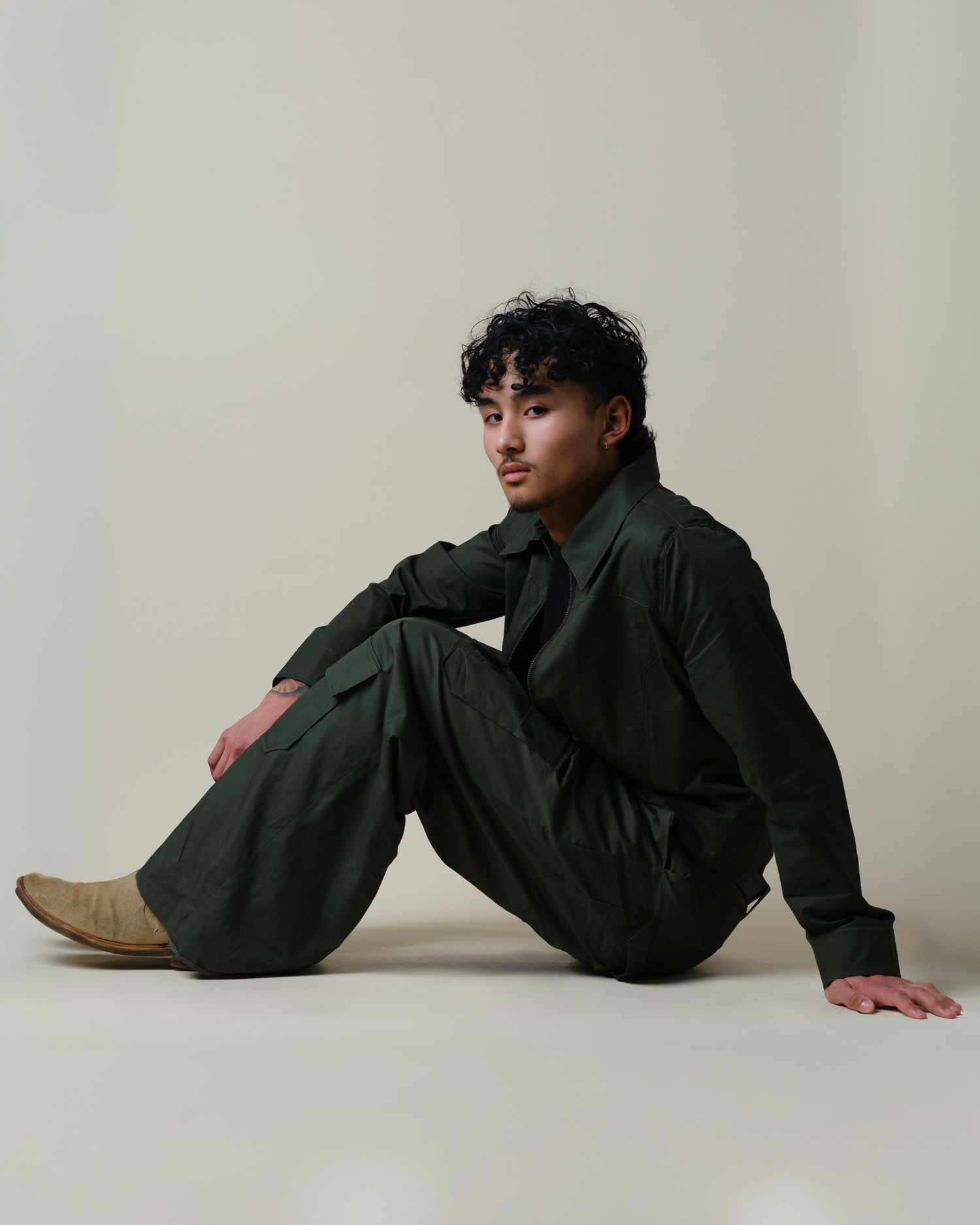 Young man in dark green cargo jumpsuit sitting on floor in minimalist studio setting.