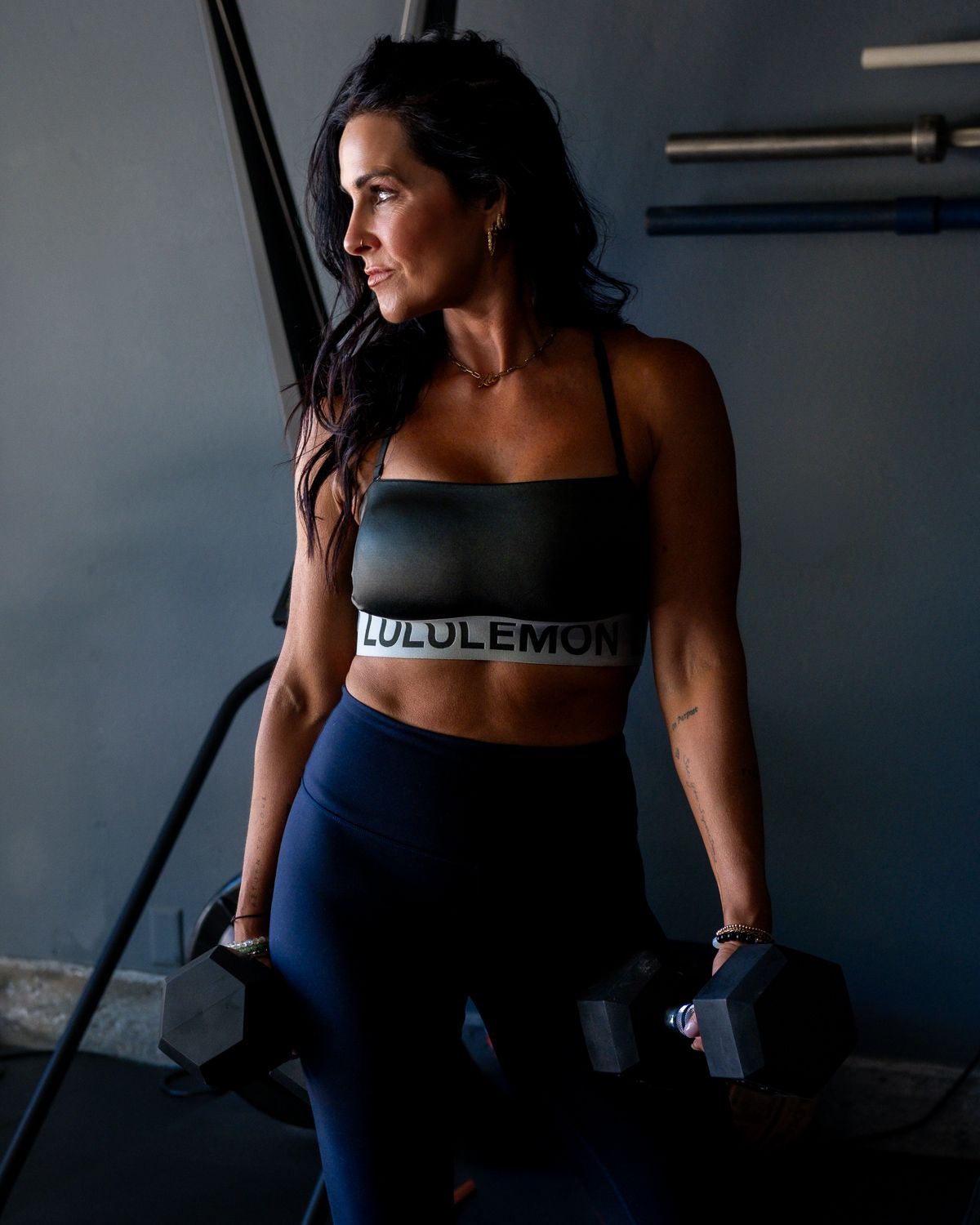 Fit woman in Lululemon sports bra holding dumbbells in a dark gym setting.