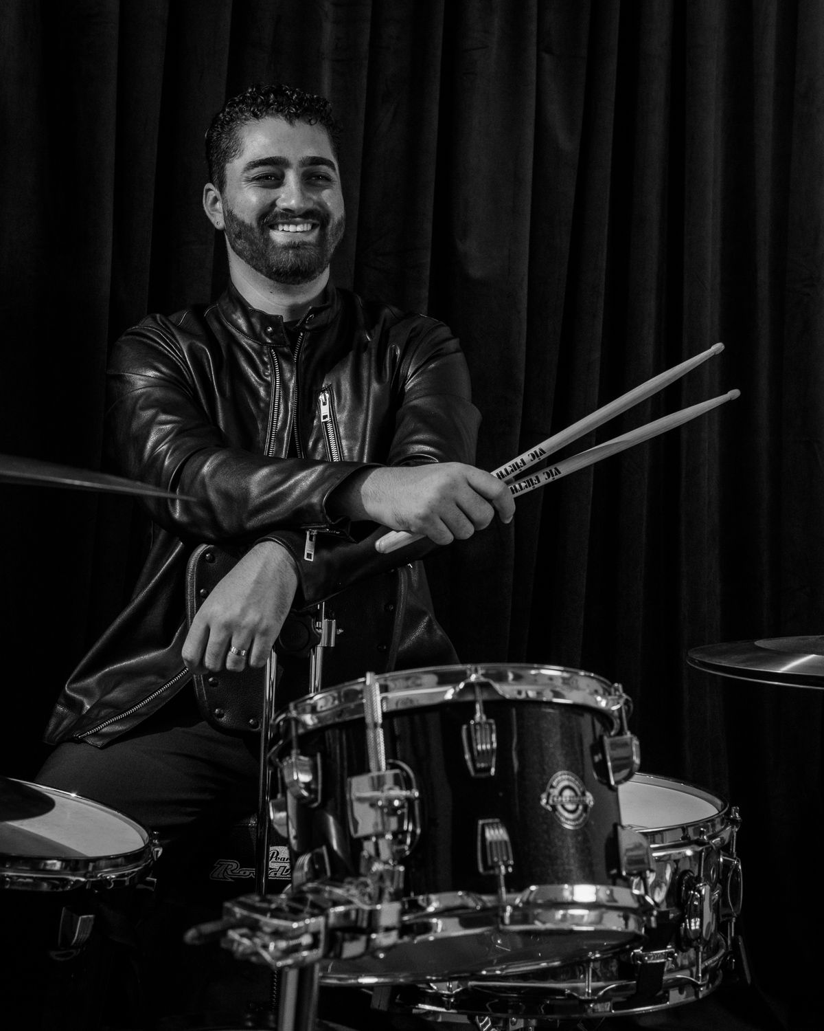 Smiling drummer holding drumsticks at a drum kit in a black and white portrait.
