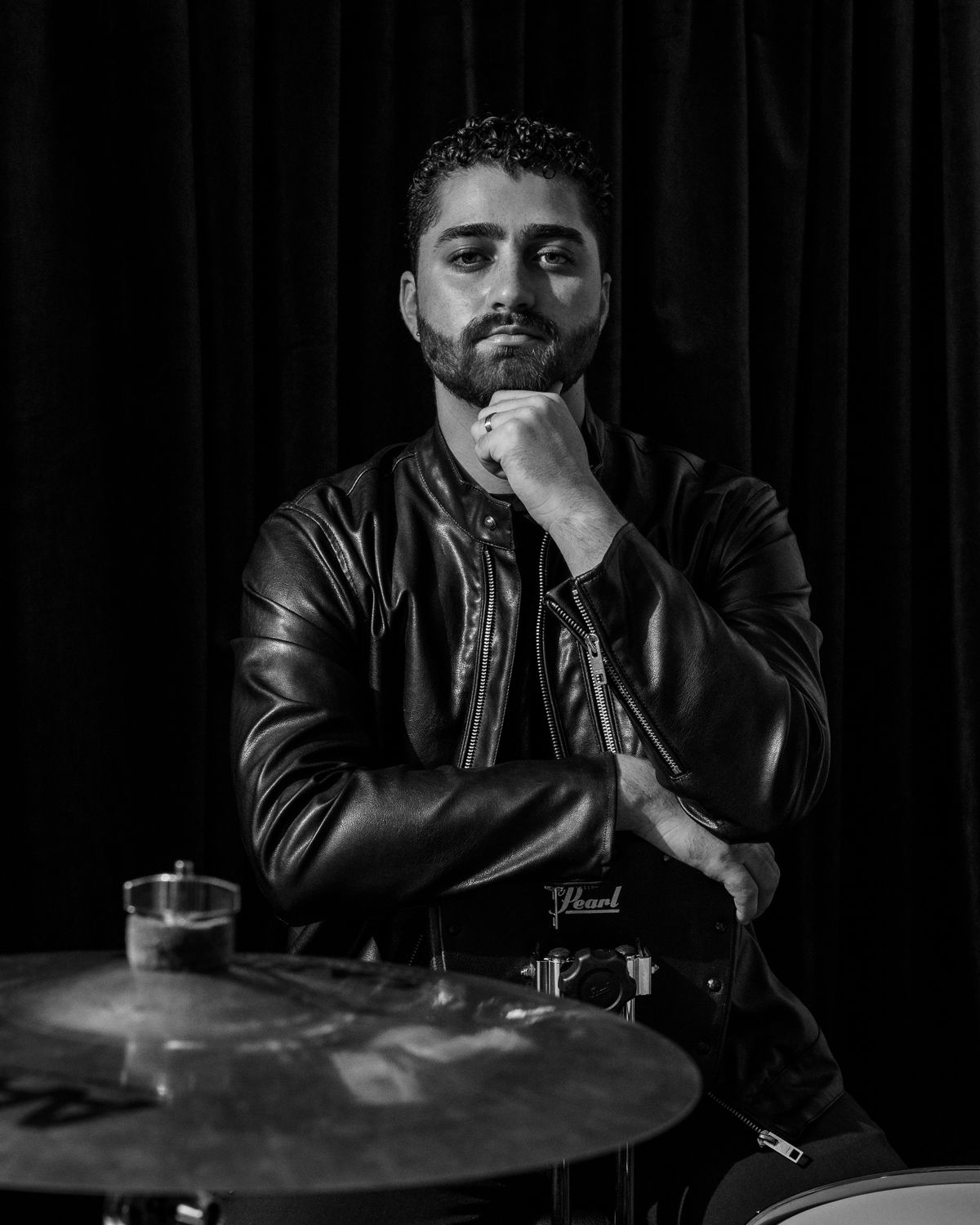 Bearded man in leather jacket posing thoughtfully beside a drum cymbal in black and white.