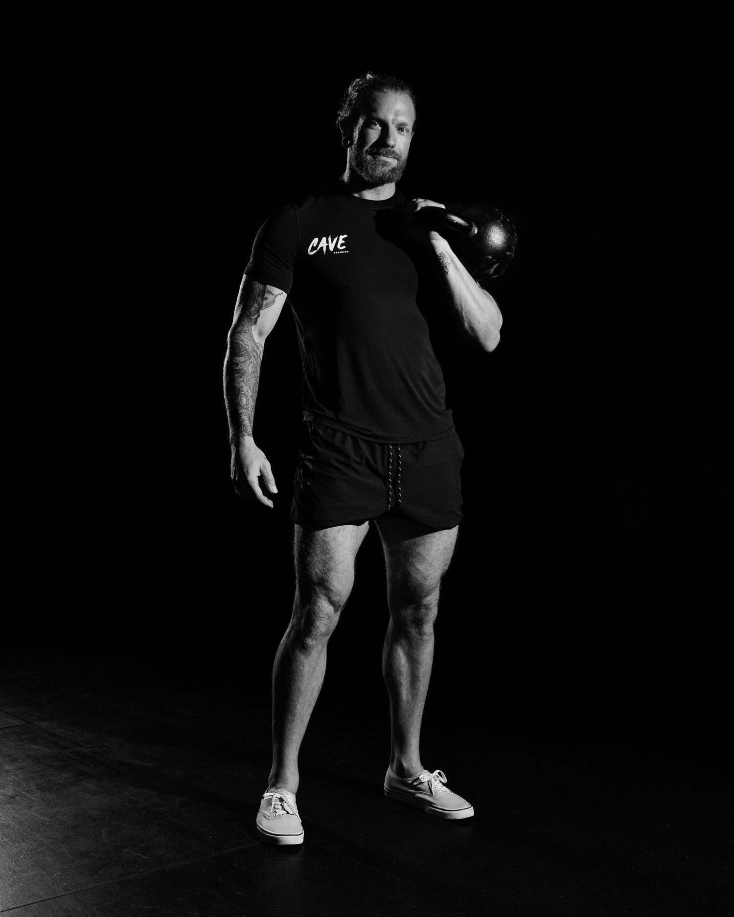 Black and white photo of a tattooed man in a Cave Training shirt holding a kettlebell.