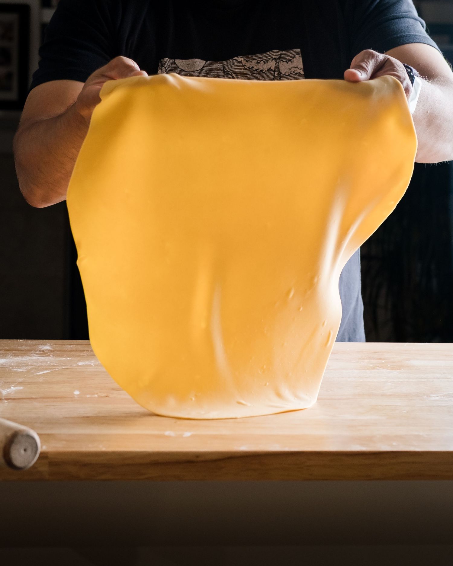 Chef stretching smooth golden fresh pasta dough above a flour-dusted wooden work surface.
