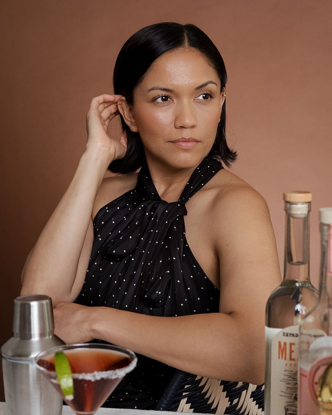 Woman in black halter top poses at bar with cocktails and tequila bottle.