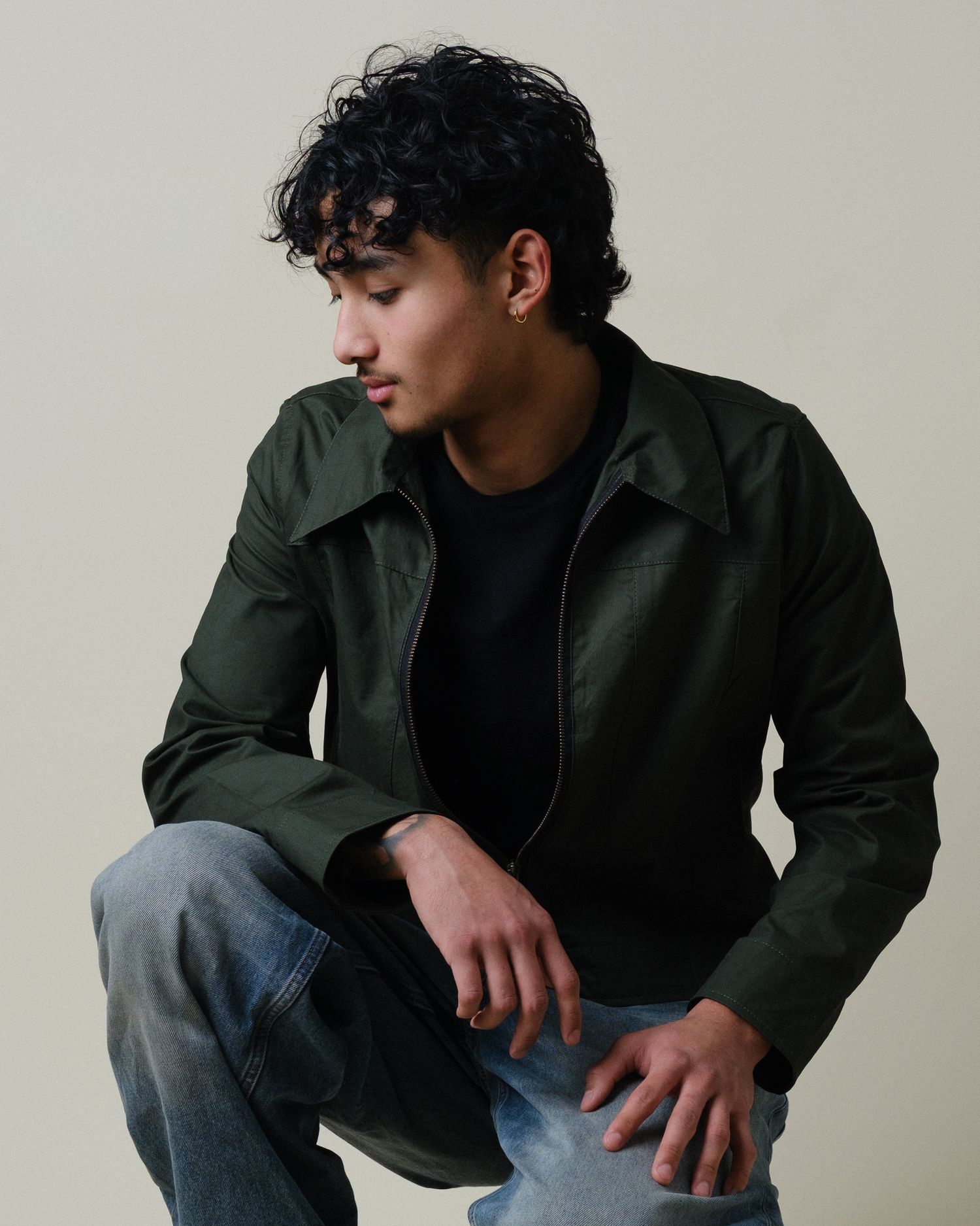 Young man in dark green jacket and jeans crouching in a neutral studio setting.