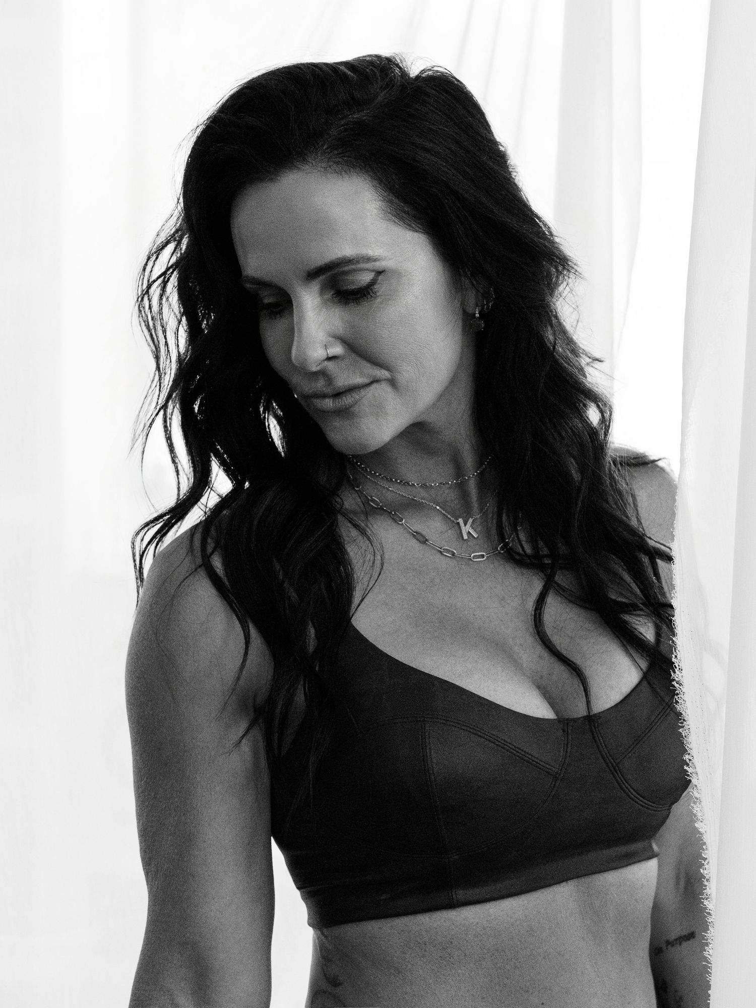 Black and white photo of a woman with dark wavy hair wearing a black sports bra and layered necklaces.