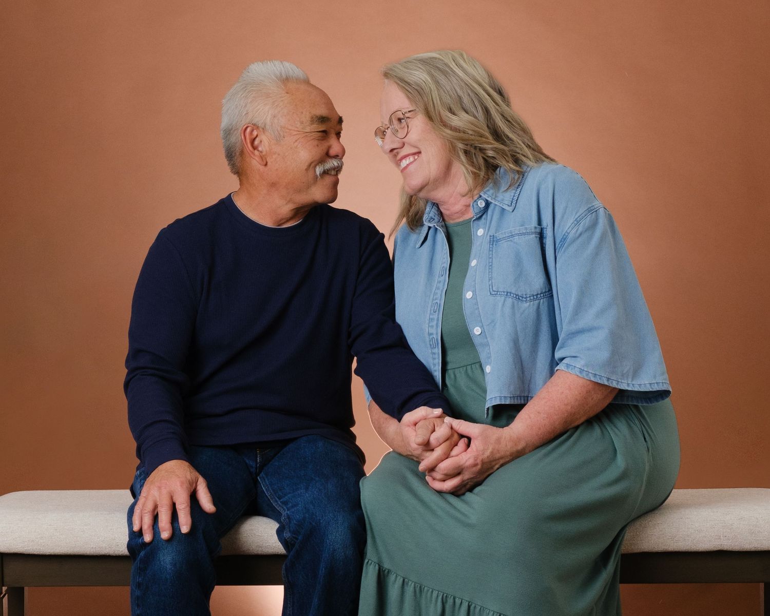 Happy senior interracial couple holding hands and smiling at each other on a bench.