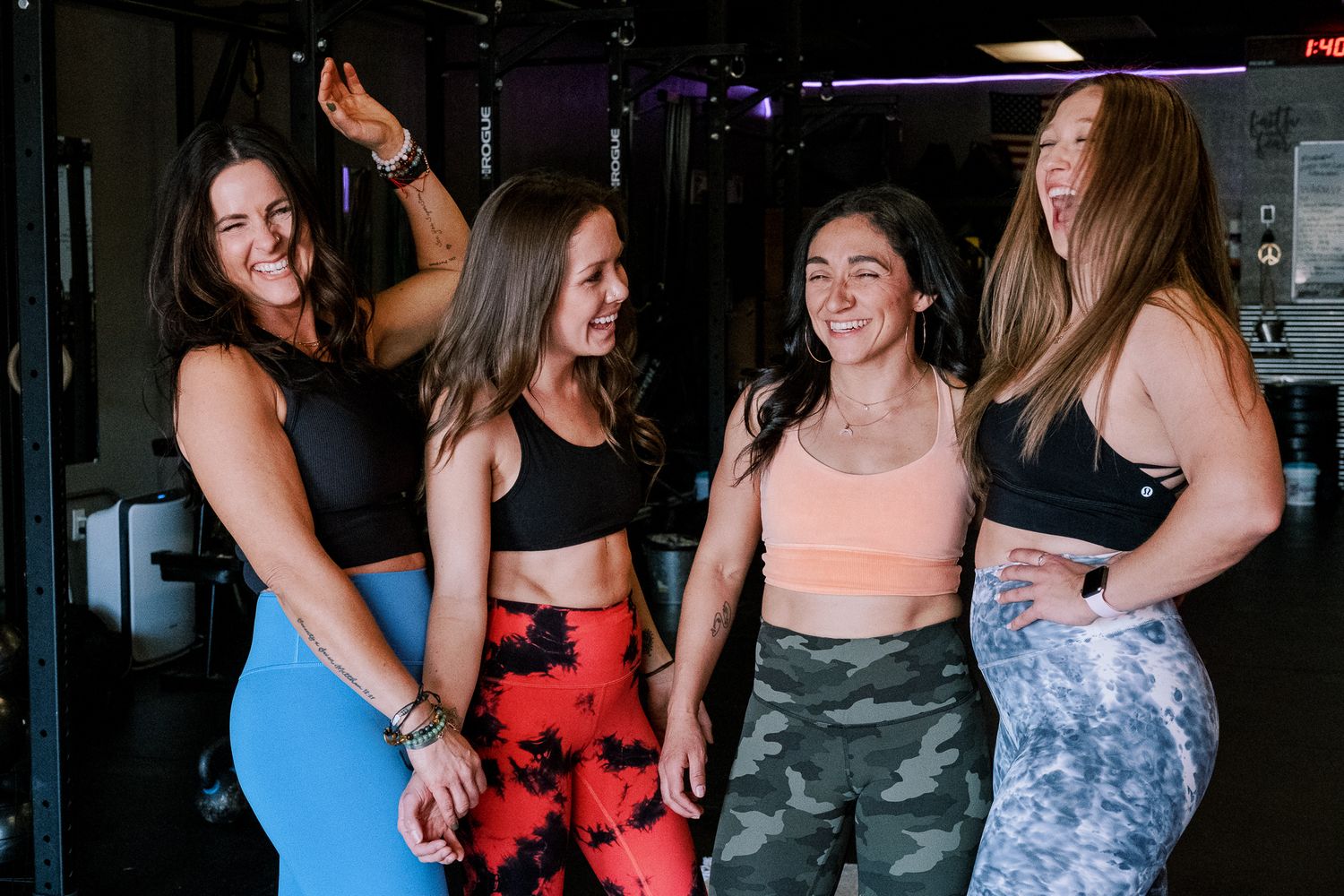 Four women in athletic wear laugh together in a gym setting wearing colorful leggings and sports bras.