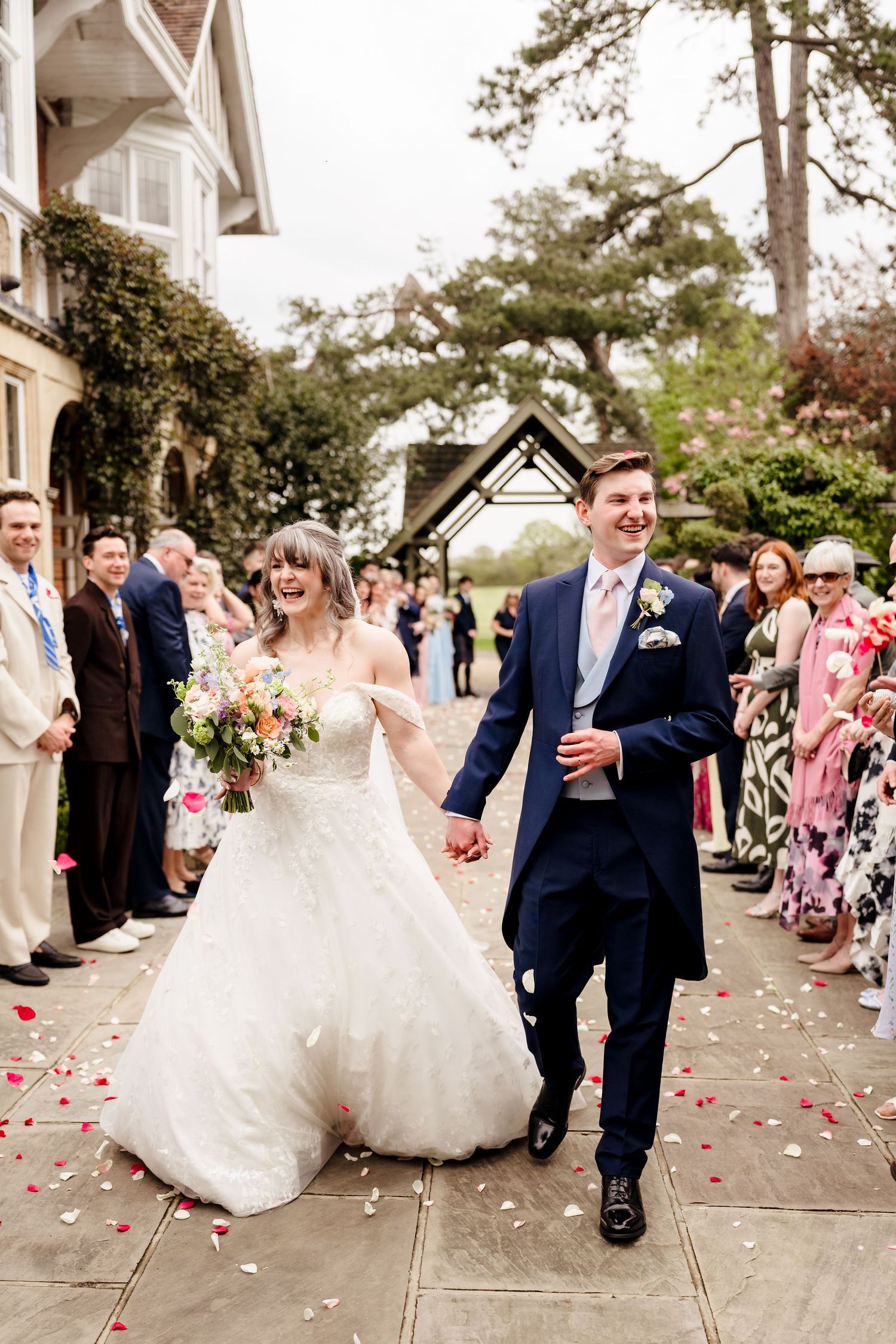Bride and groom walk hand-in-hand through rose petal confetti outside elegant wedding venue.