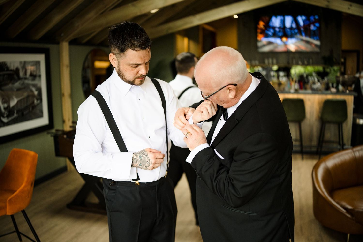 Groom with tattoos and suspenders gets help with cufflinks from older man in tuxedo at wedding venue bar.