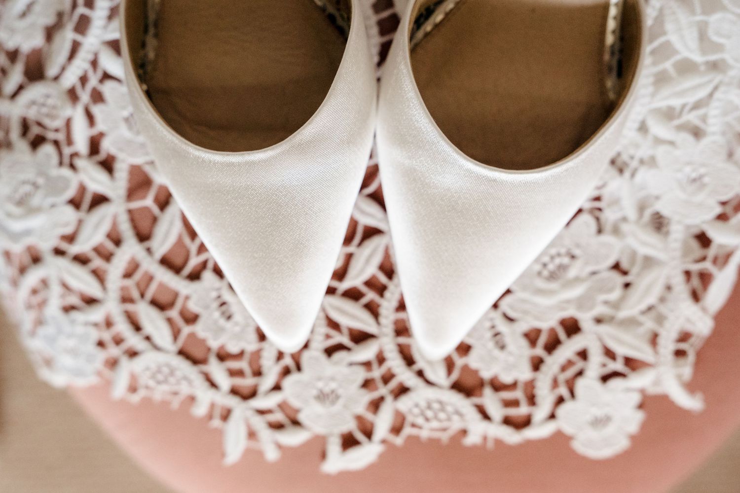 White satin pointed-toe bridal heels resting on delicate lace wedding dress fabric.