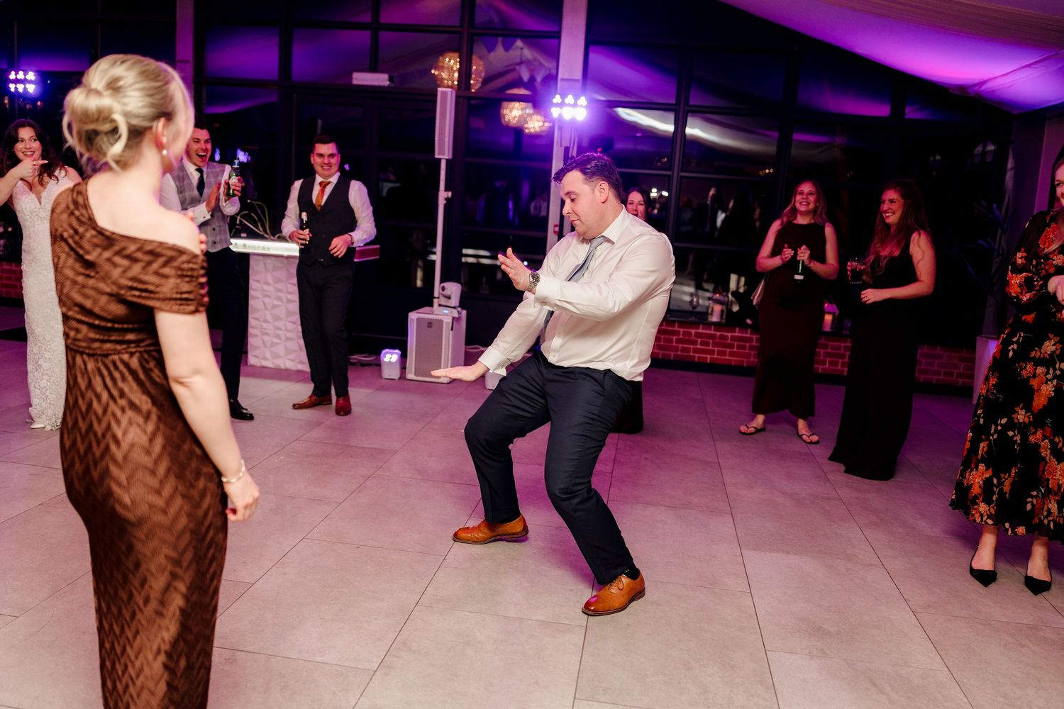 Wedding guest energetically dancing and crouching on the dance floor while guests watch and cheer at reception.