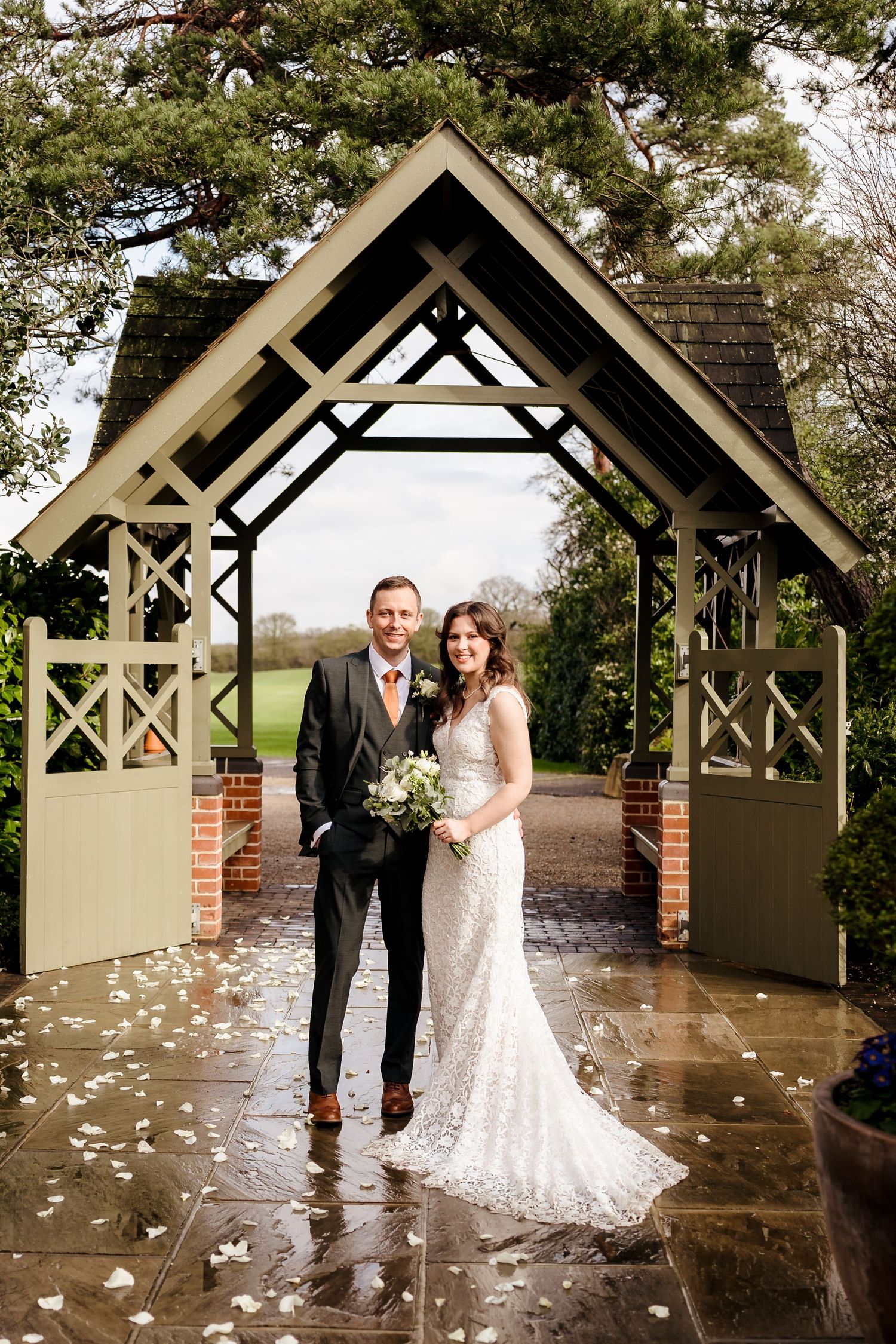 Bride and groom posing under wooden arch gateway surrounded by white rose petals on wet stone path.