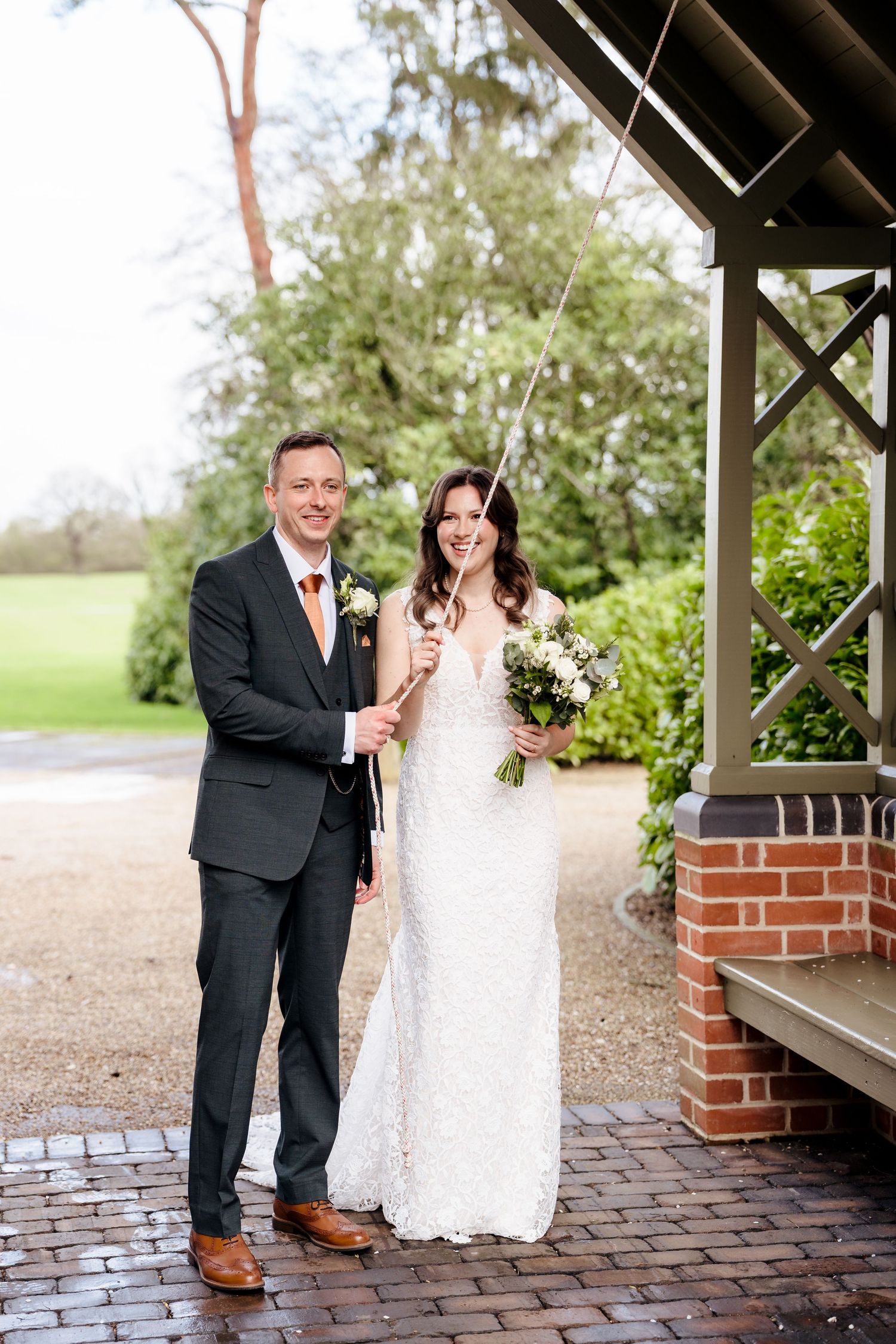 A smiling bride in a white lace gown and groom in a dark suit pose with sparklers at an outdoor wedding venue.