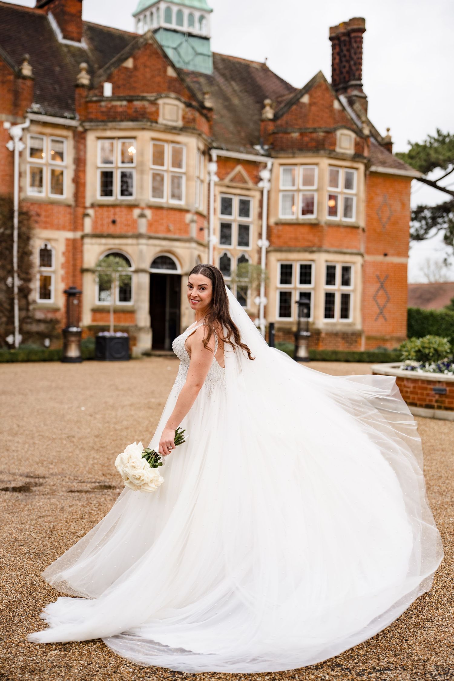 Bride in flowing white ballgown twirling with bouquet in front of a grand red brick manor house.