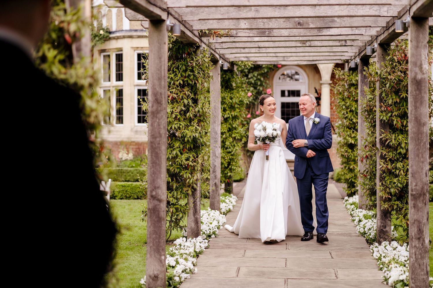 Bride walks down outdoor aisle with escort under pergola surrounded by lush greenery at elegant wedding venue.
