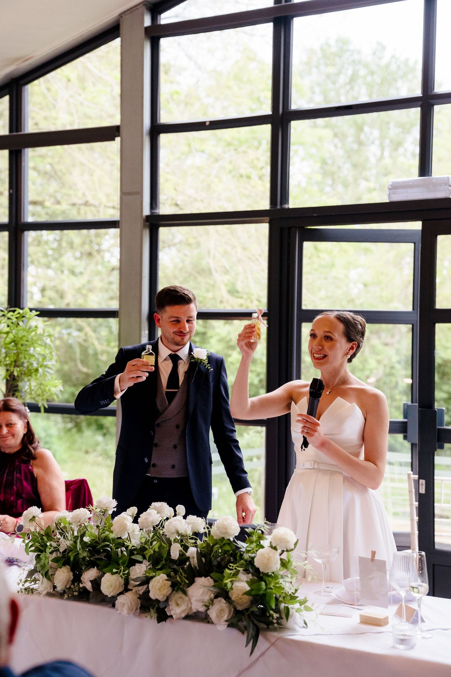 Bride speaking into microphone while groom raises glass during wedding reception speech at floral head table.