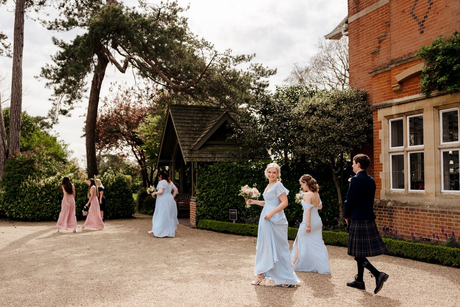 Bridesmaids in light blue gowns and pink dresses walking along gravel path beside elegant red brick wedding venue.