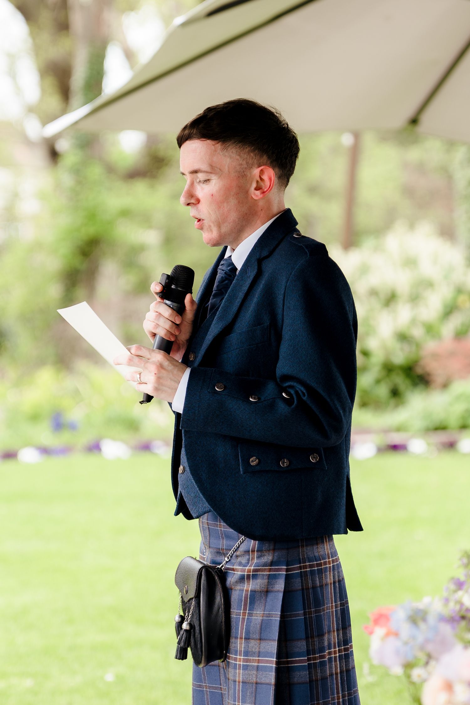 Young man in a navy jacket and tartan kilt reading a speech into a microphone at an outdoor wedding.