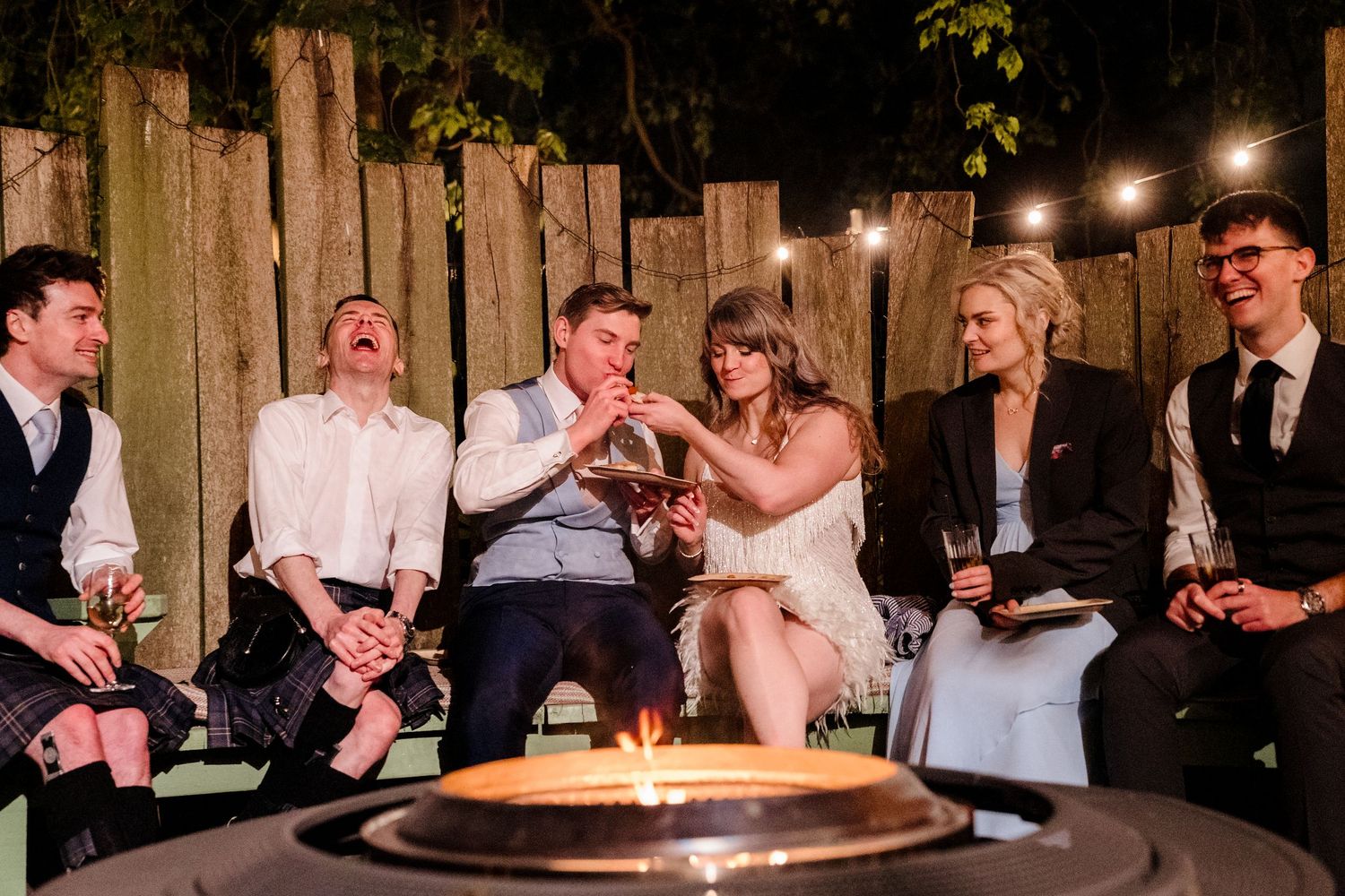 Wedding guests laughing and socializing around a glowing fire pit under string lights at an outdoor reception.