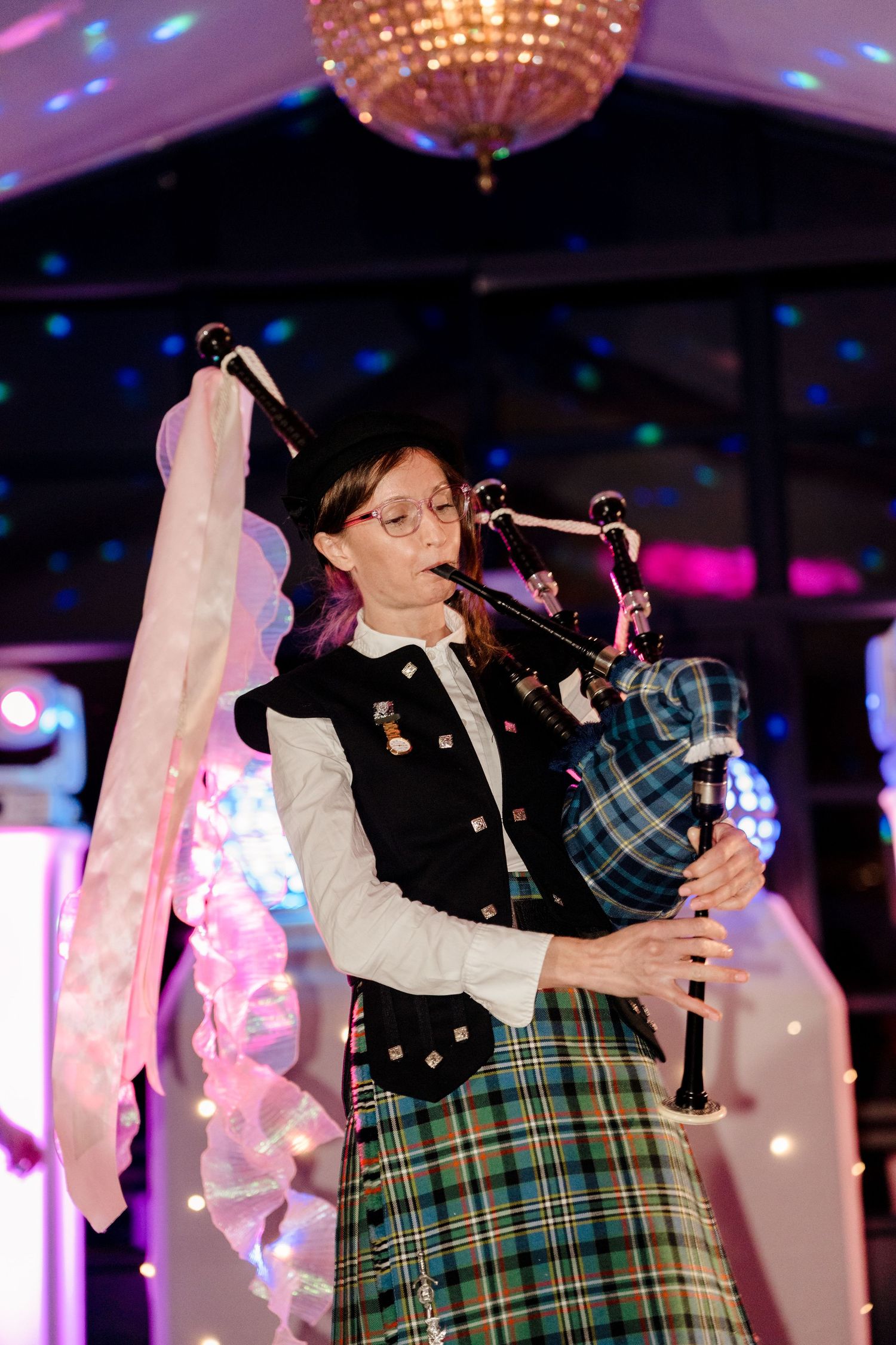 Female bagpiper in traditional Scottish tartan kilt performing under a crystal chandelier at a wedding reception.