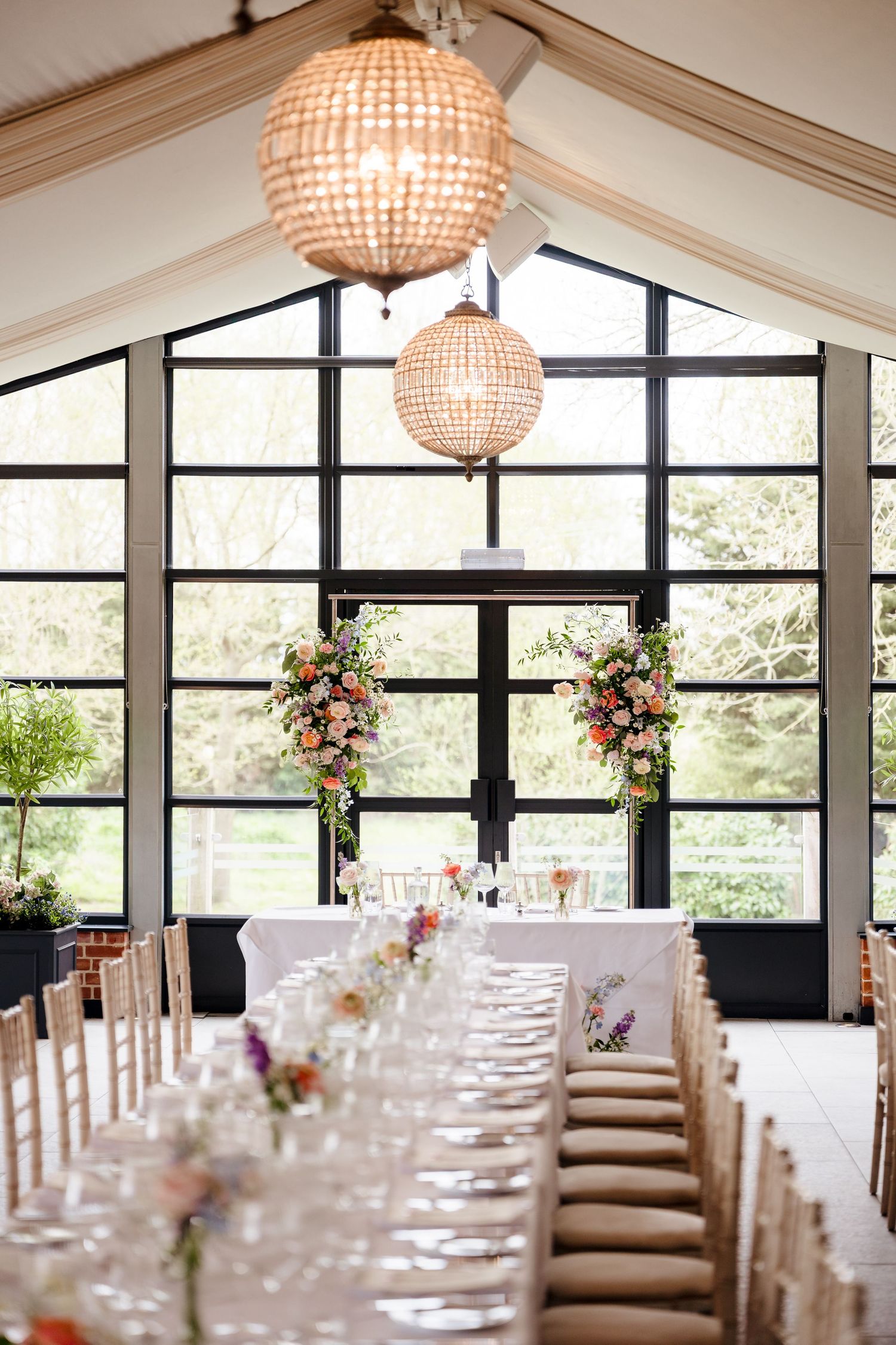 Bright wedding reception hall with long dining table, floral arrangements, and beaded chandeliers.