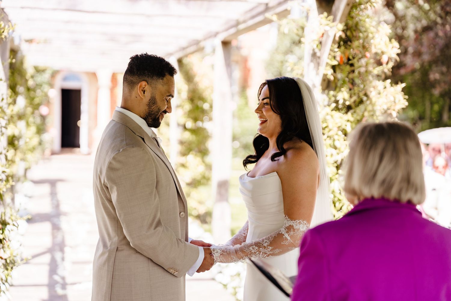 Bride and groom exchange vows outdoors under floral arch with officiant during wedding ceremony.