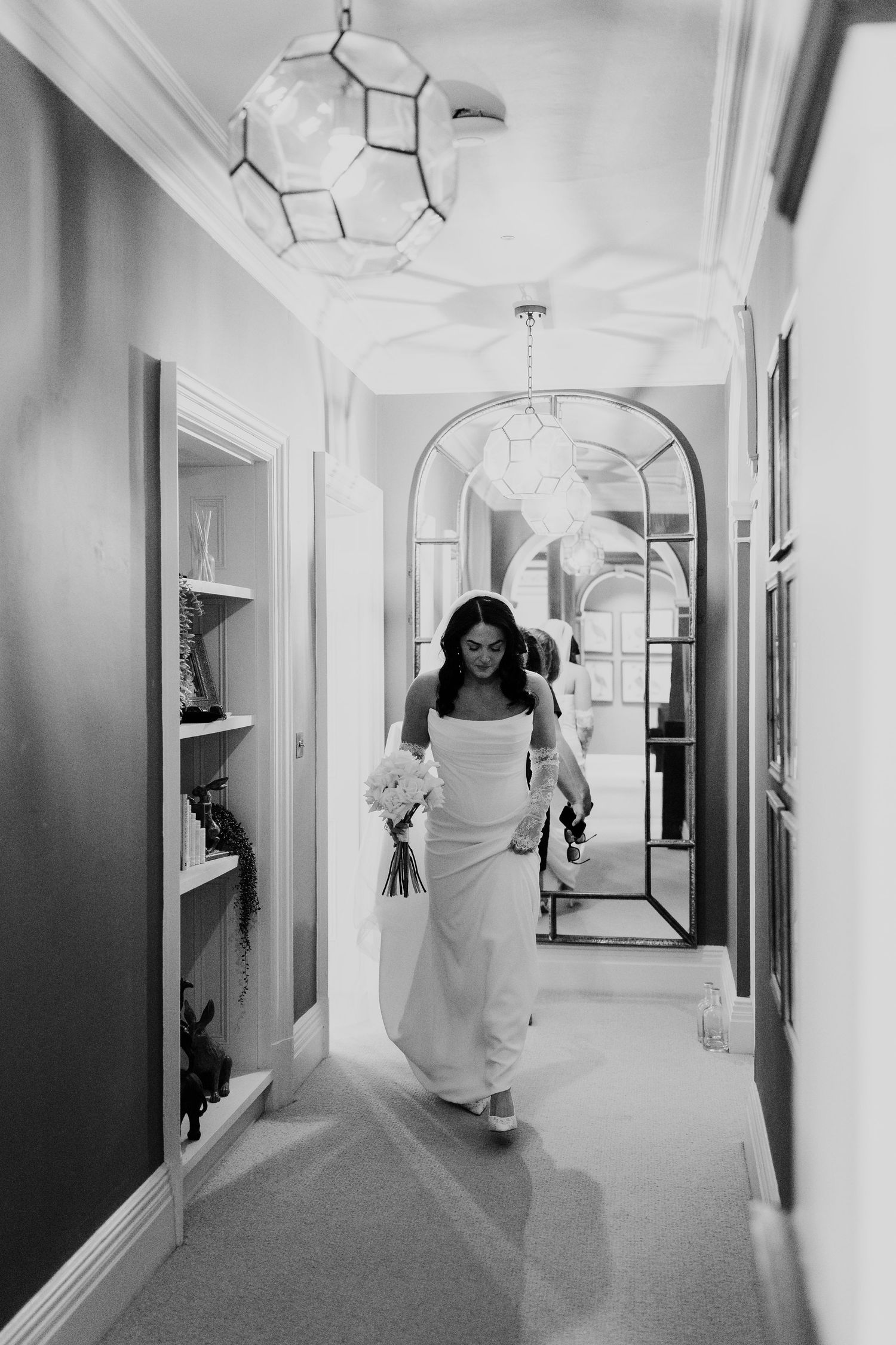 Bride in white gown walking down elegant hallway with chandelier lighting.