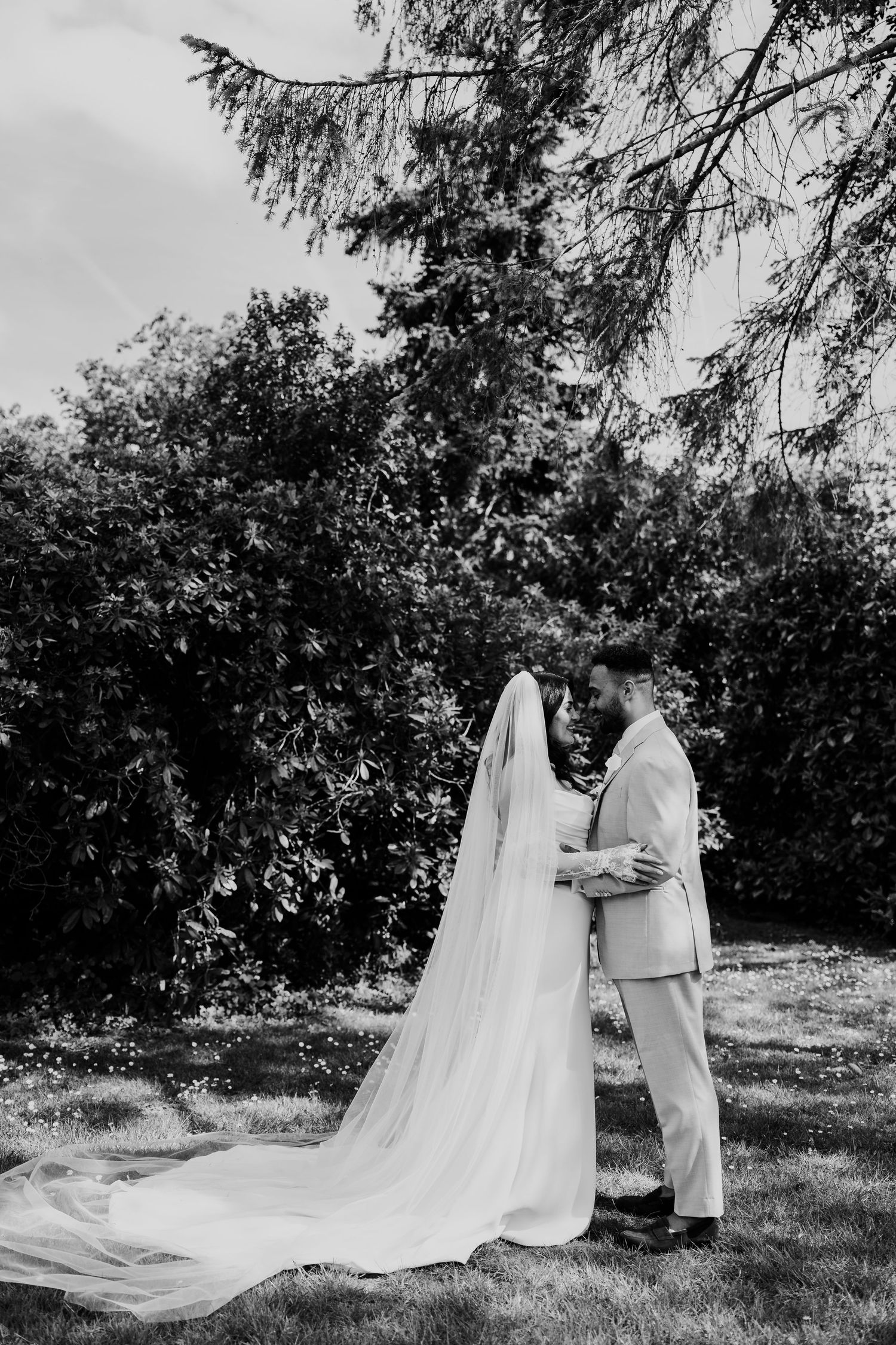 Black and white romantic portrait of bride and groom embracing in garden with flowing cathedral veil.