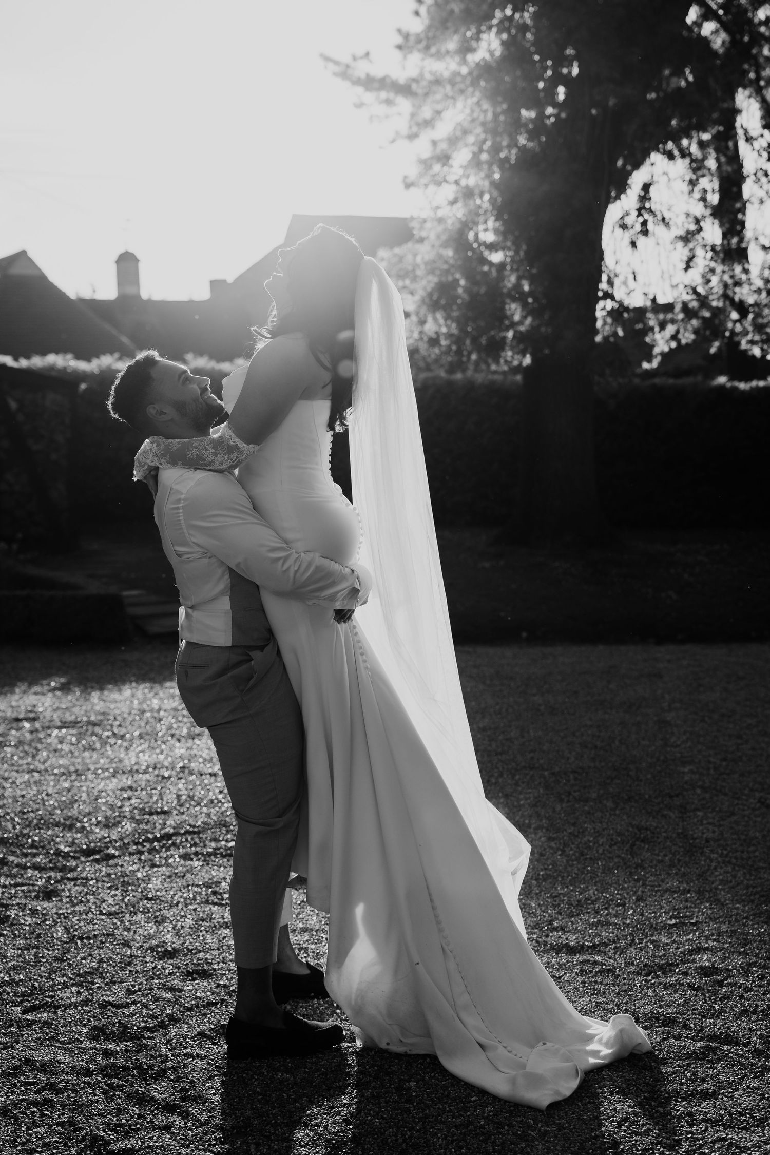 Romantic black and white photo of groom lifting bride in flowing veil at golden hour.