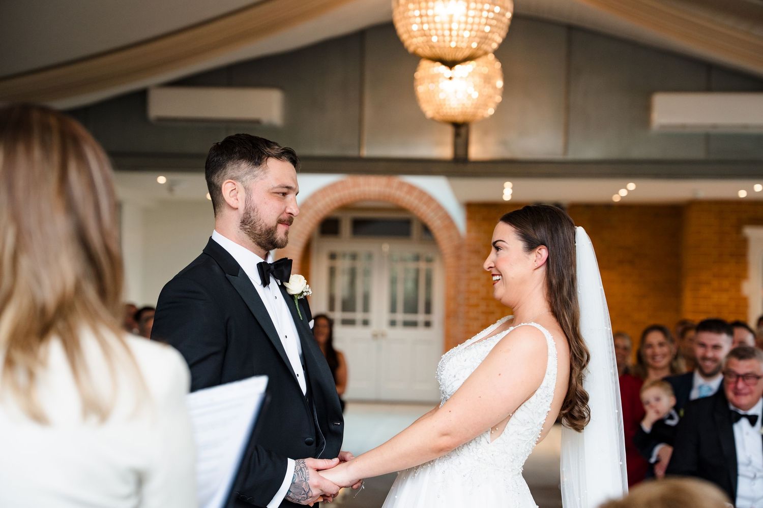 Bride and groom holding hands and smiling during wedding ceremony in elegant venue with chandelier.