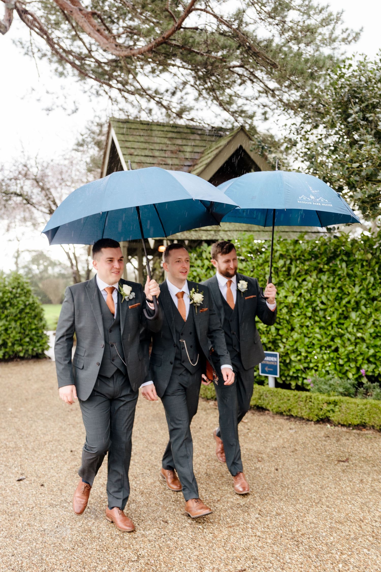 Three groomsmen in grey suits walk with blue umbrellas on a rainy wedding day.