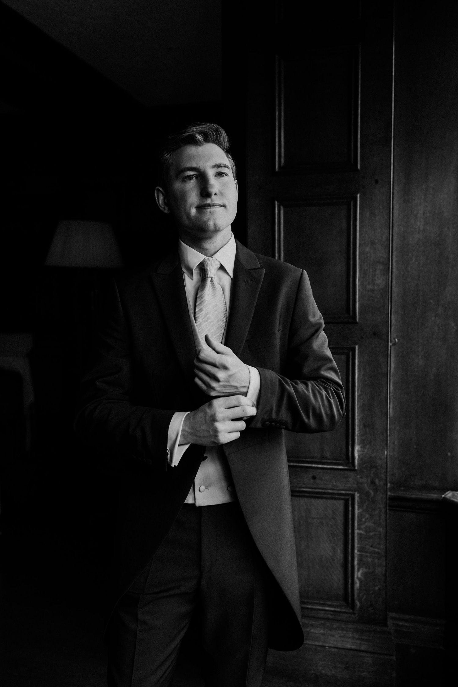 Dapper groom in dark suit adjusting cufflinks in black and white portrait by wooden door.