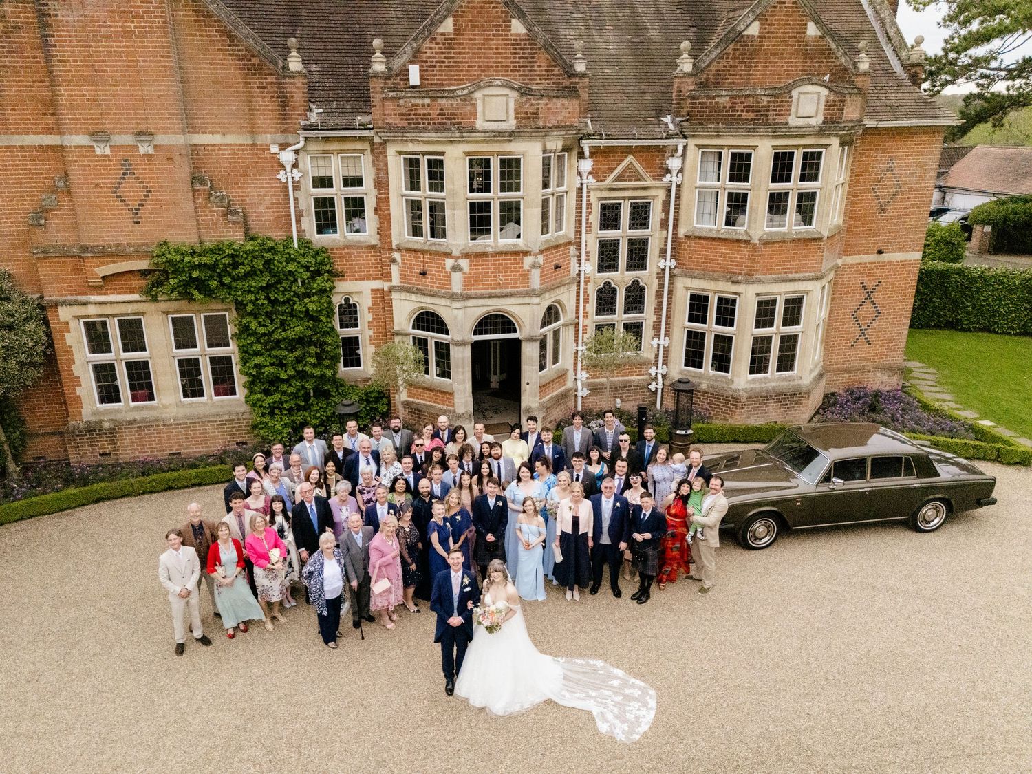 Aerial view of wedding party and guests posing outside a grand brick manor house with a vintage car.