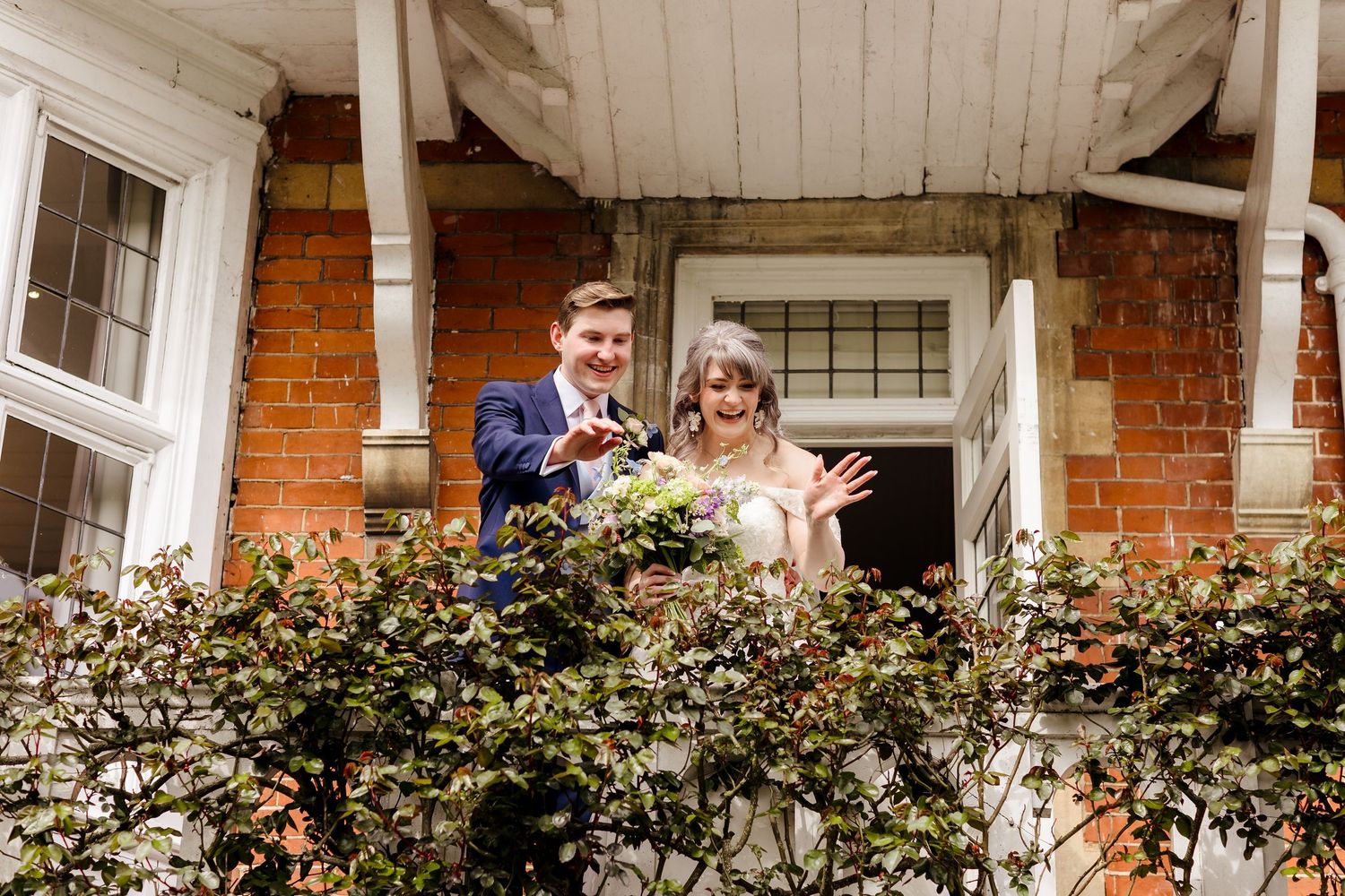 Newlywed couple waving joyfully from a brick building balcony surrounded by green foliage.