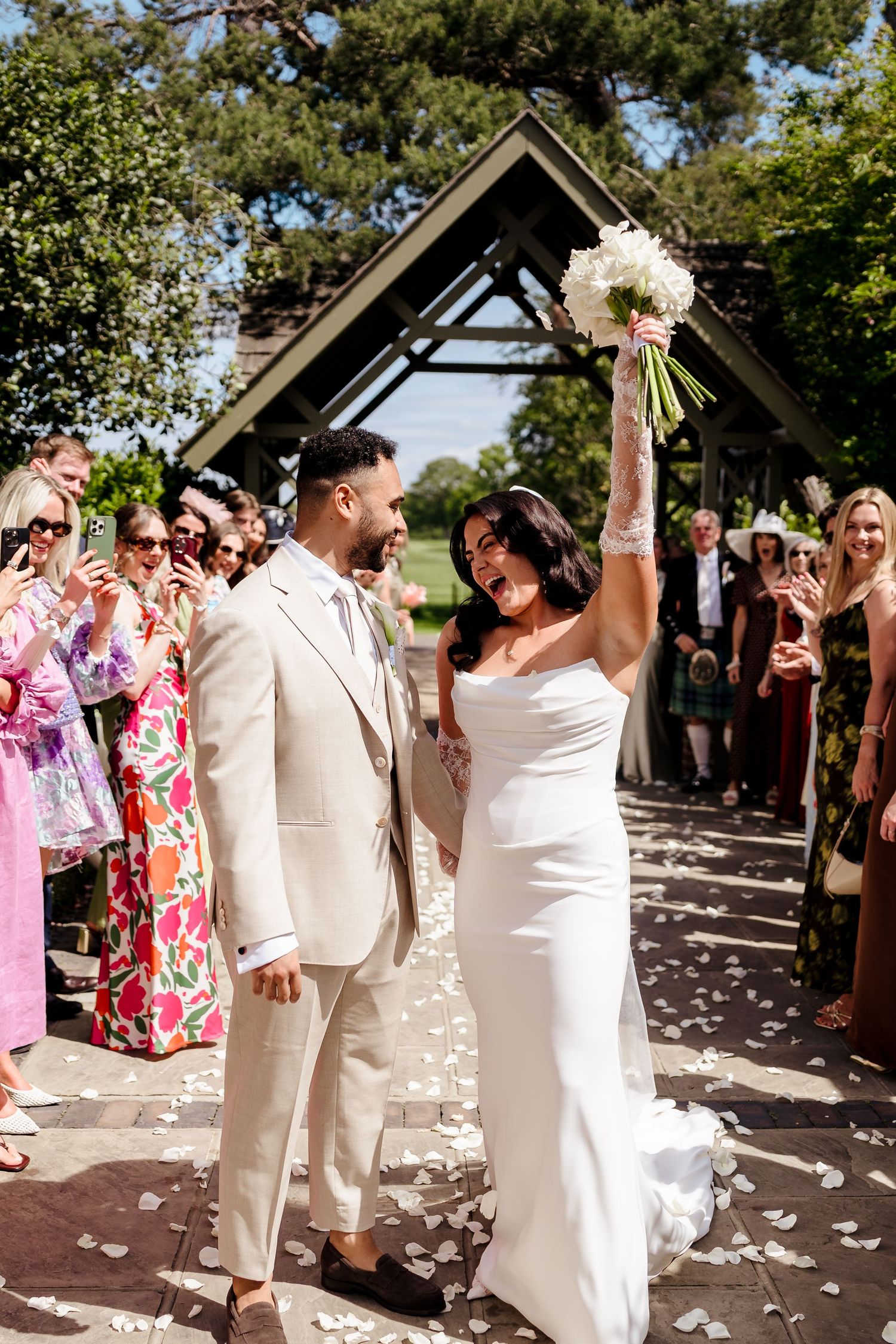 Joyful bride raises white bouquet while walking with groom in beige suit through confetti-lined aisle.
