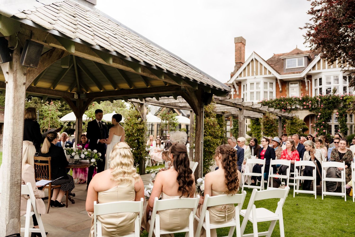 Outdoor wedding ceremony under wooden pergola with bridesmaids seated in white chairs before historic manor.