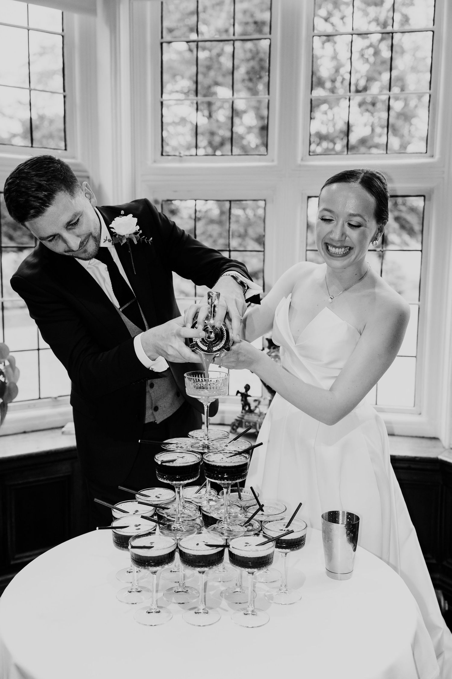 Black and white photo of laughing bride and groom pouring champagne tower at wedding reception.