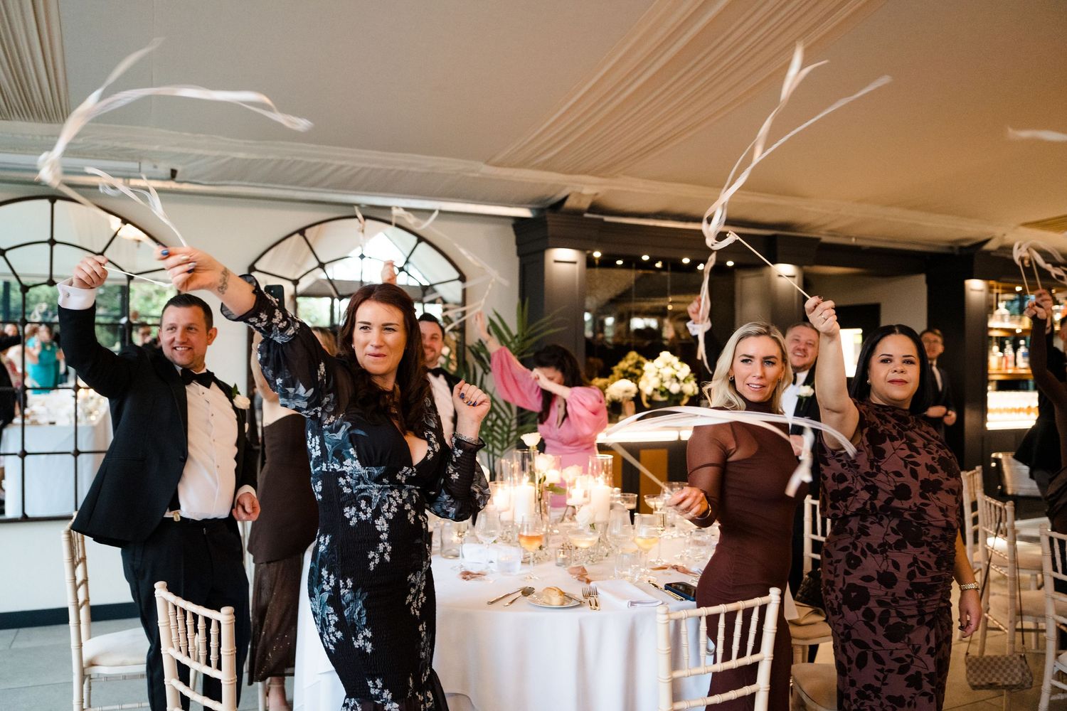 Wedding guests celebrate joyfully waving ribbon wands at a decorated reception dinner table.