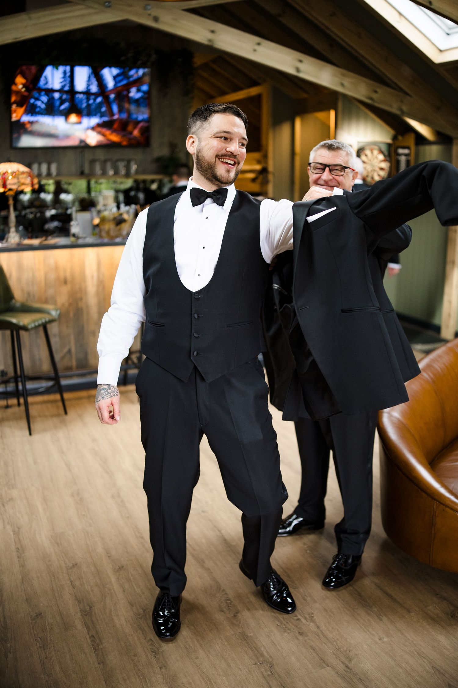 Smiling groom in black tuxedo vest being helped into his jacket by an older man in a stylish bar setting.