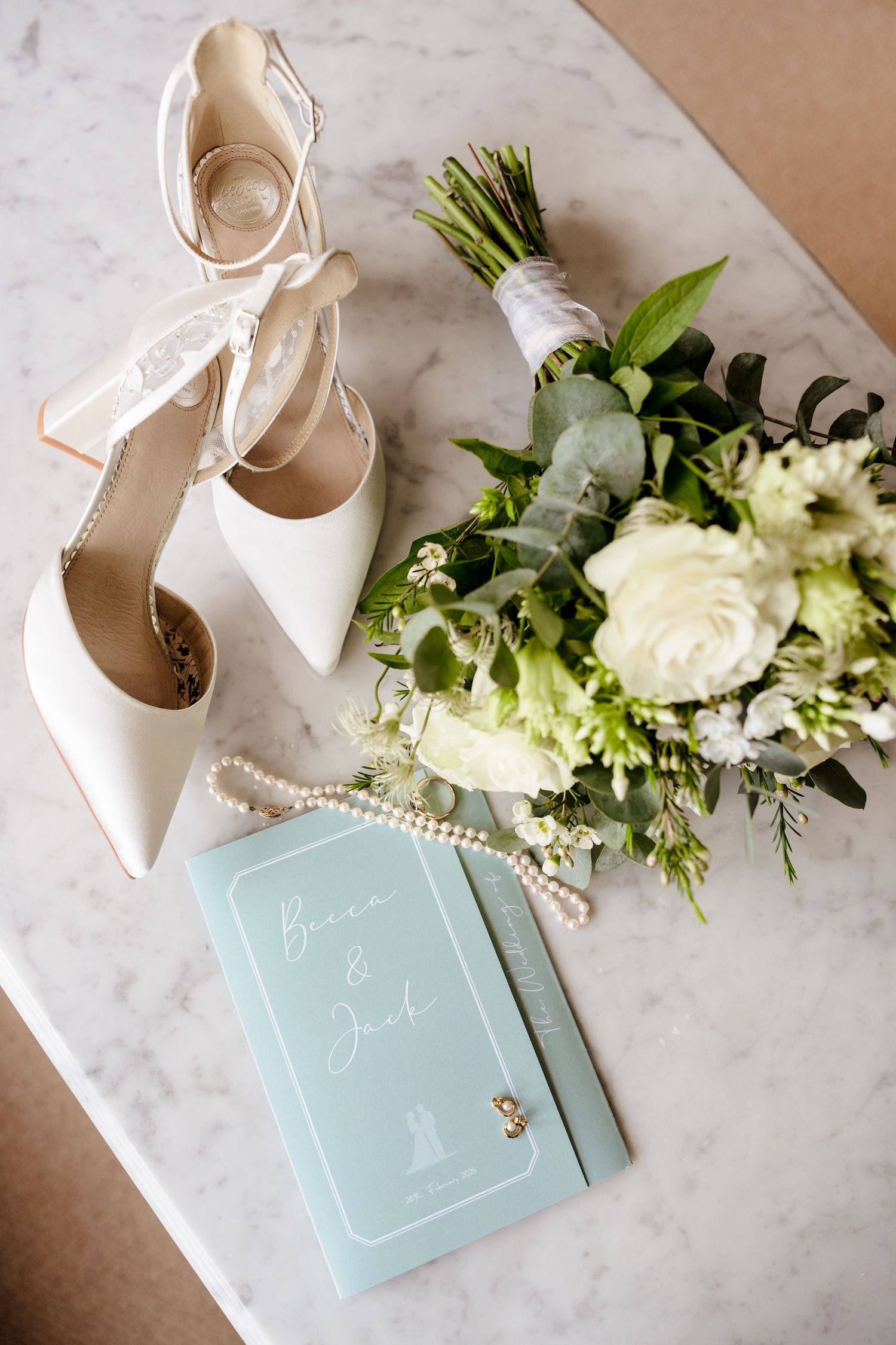 Elegant wedding flat lay featuring white heels, pearl necklace, floral bouquet, and mint green invitation on marble.