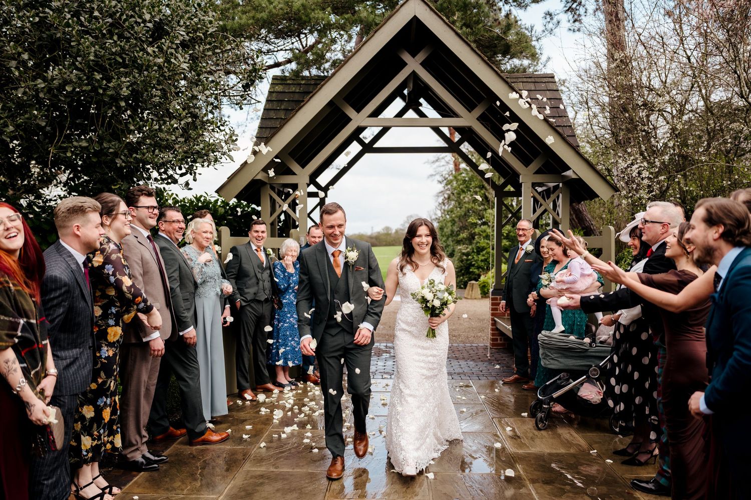 Newlyweds walk through confetti shower under wooden pergola after rainy wedding ceremony.