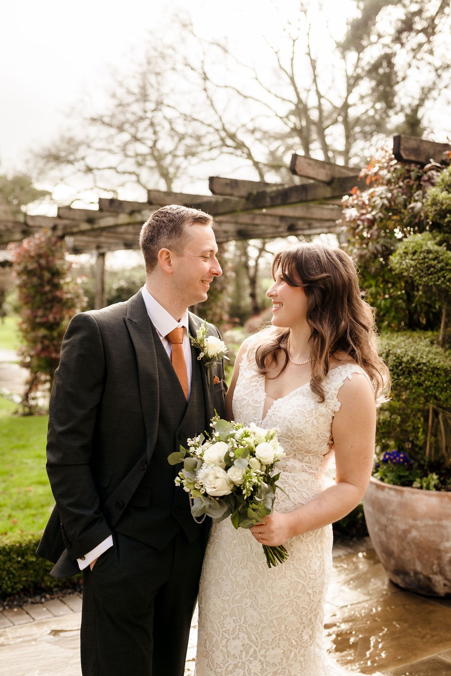 Bride and groom smiling at each other in a garden, bride holding white bouquet, groom in black suit with orange tie.