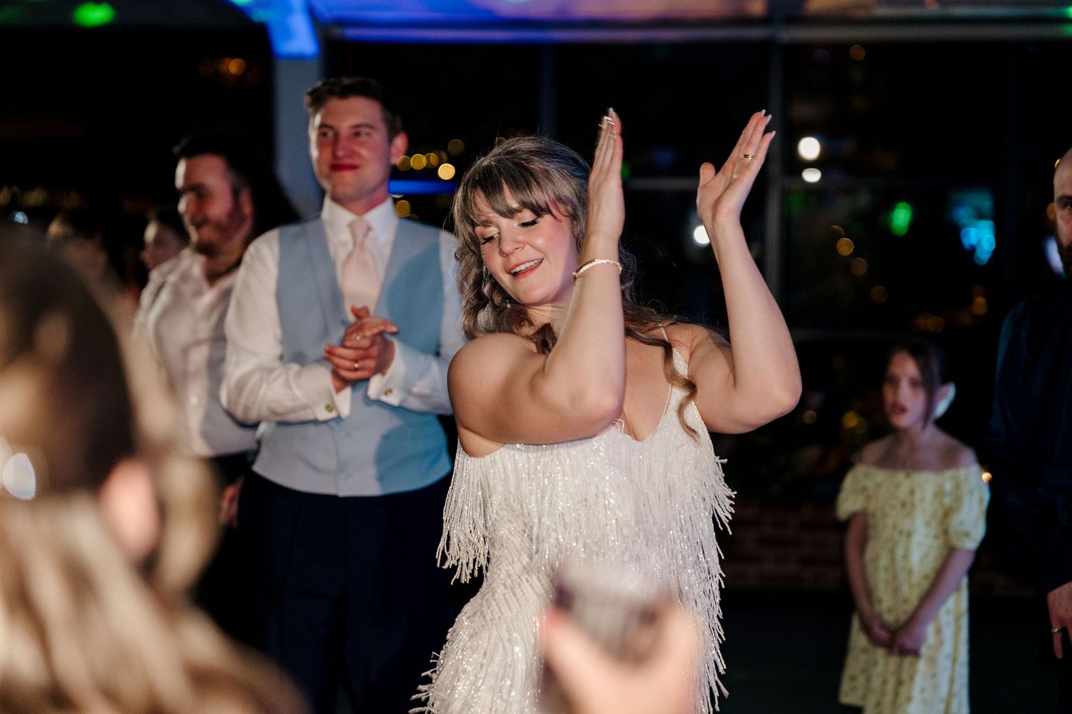 Bride in a white fringed dress dancing joyfully with raised hands at her wedding reception celebration.