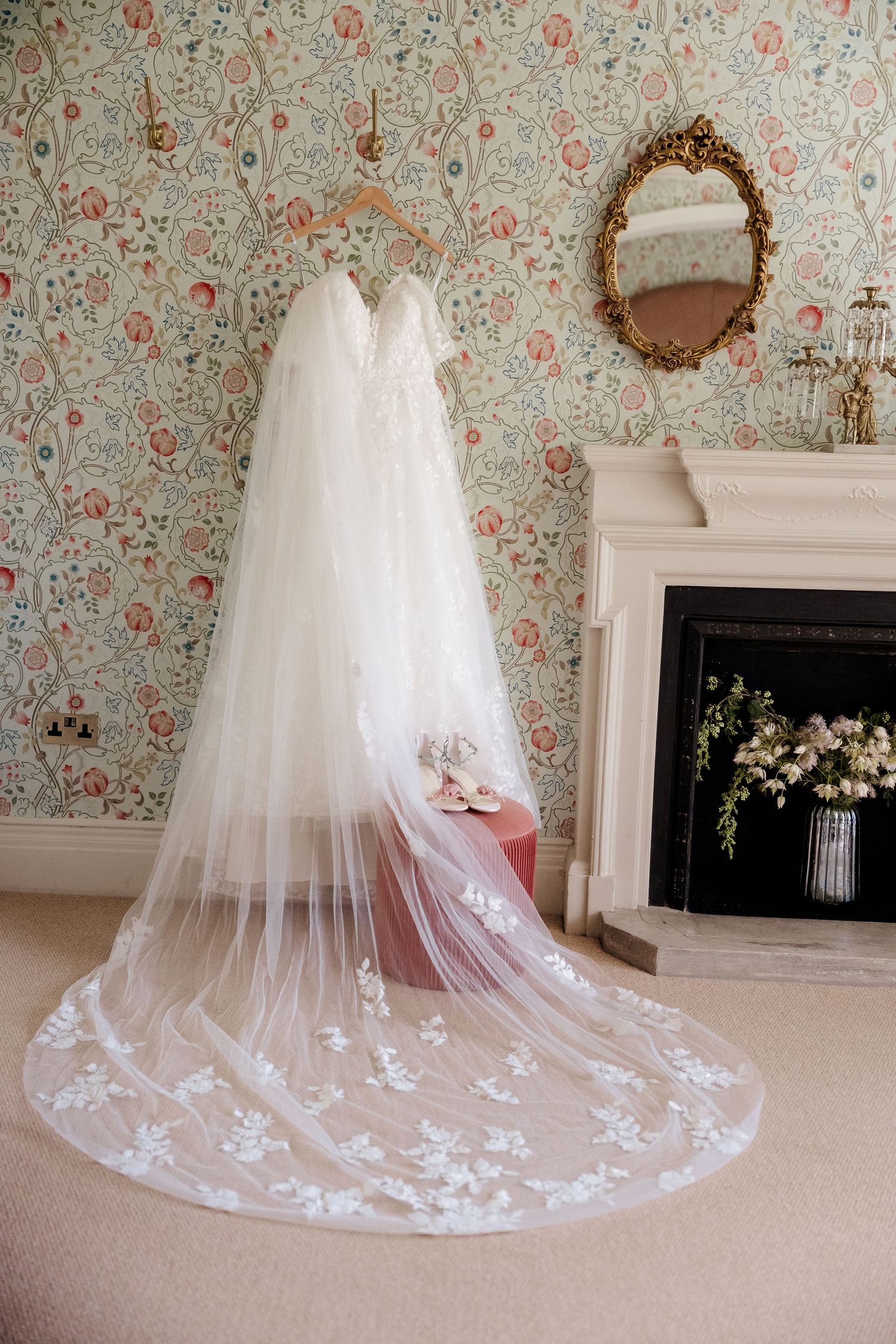 Elegant wedding dress with floral lace veil displayed on hanger in vintage room with floral wallpaper and ornate fireplace.